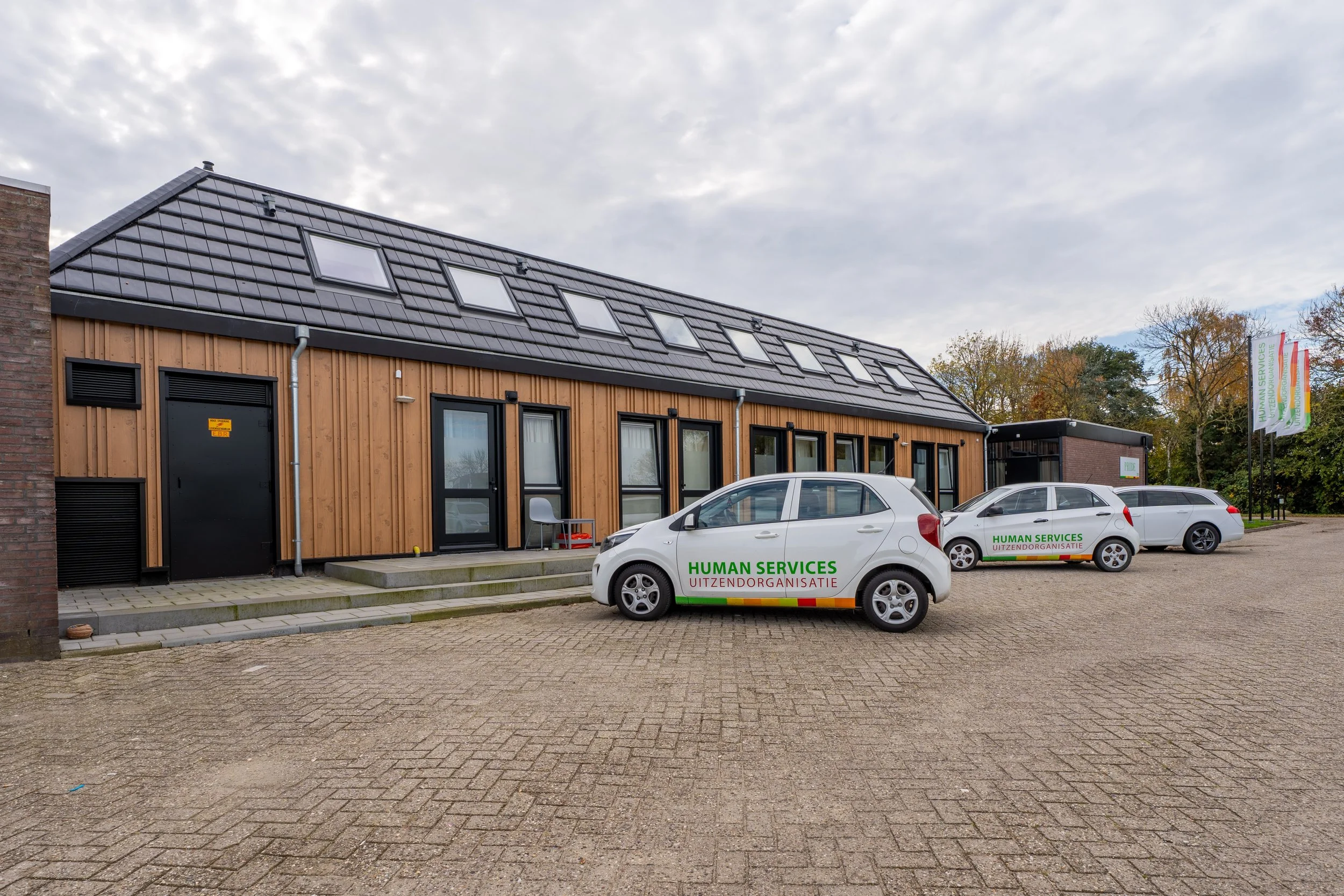 A modern building with wooden siding and a sloped roof with multiple skylights, parked with three cars in front that have 'Human Services Uitzendorganisatie' written on them, and trees with autumn foliage in the background.