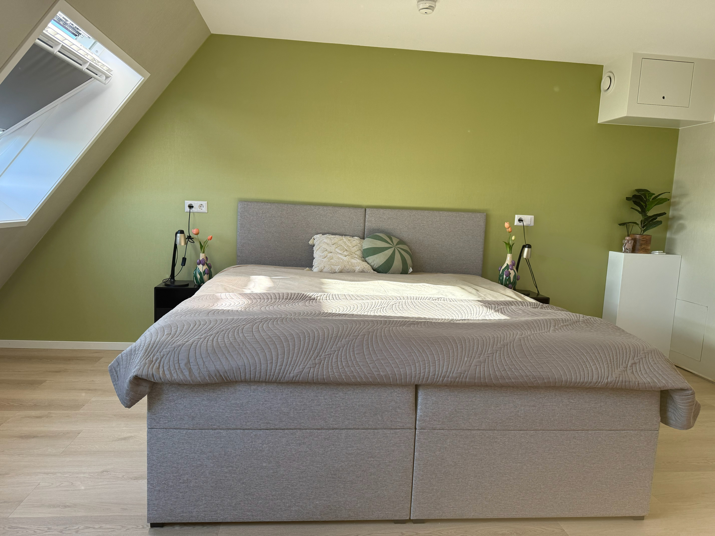 A bedroom with a large bed, gray headboard, beige quilt, and decorative pillows, set against a green accent wall. There are black nightstands with lamps and vases with flowers on either side of the bed, a white cabinet with potted plant, and a window