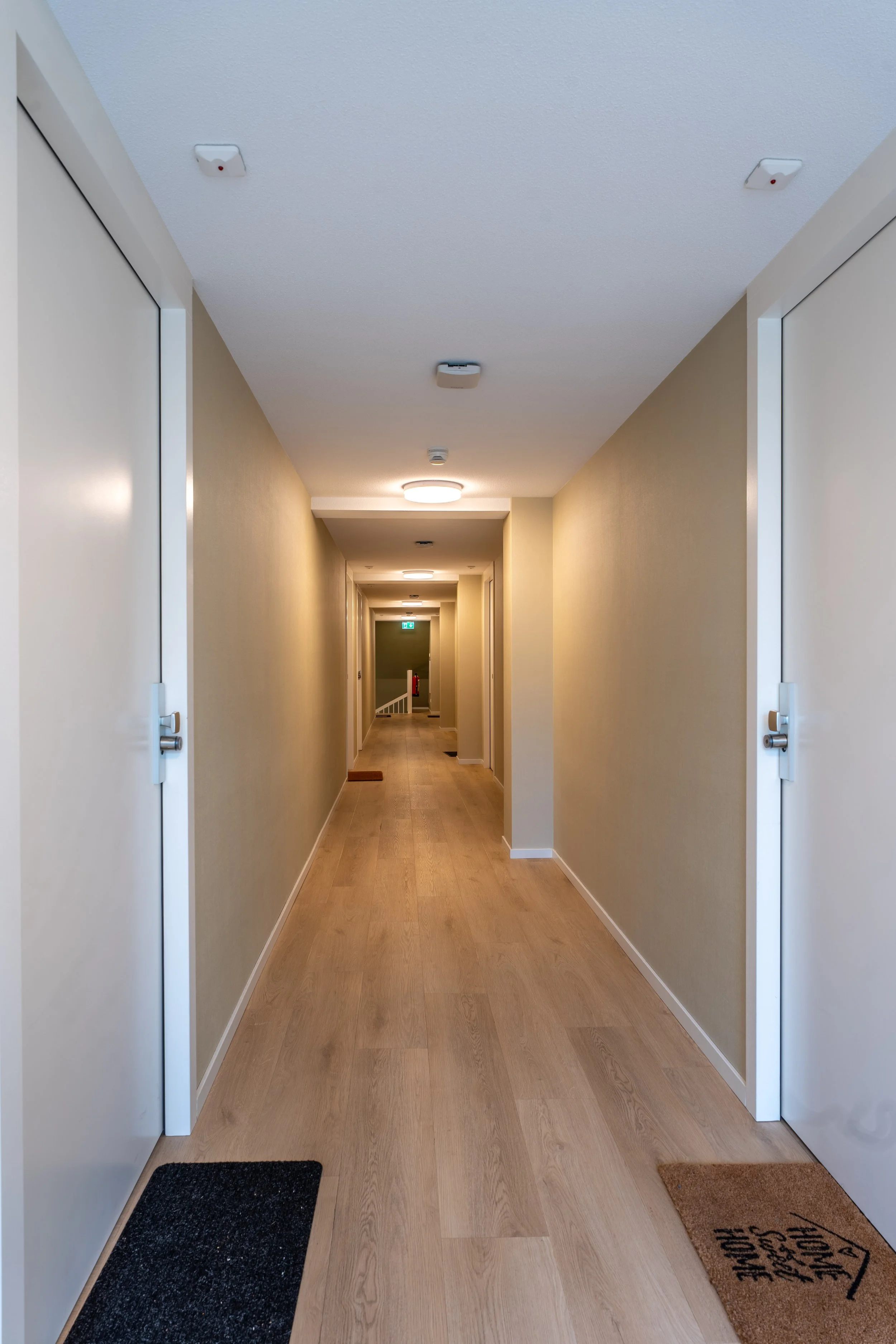 A clean, well-lit corridor with closed white doors on each side, light-colored wooden flooring, and a few wall-mounted smoke detectors and ceiling lights.