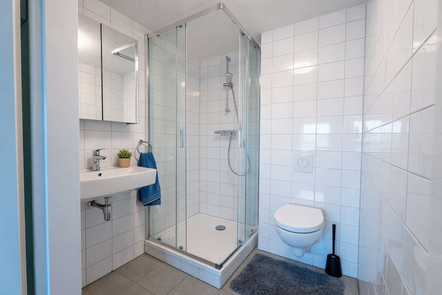 A small bathroom with a shower enclosure, a wall-mounted toilet, a white sink with a blue towel hanging, a mirror cabinet, and a small potted plant on the sink. There is a gray bath mat and a black toilet brush holder.