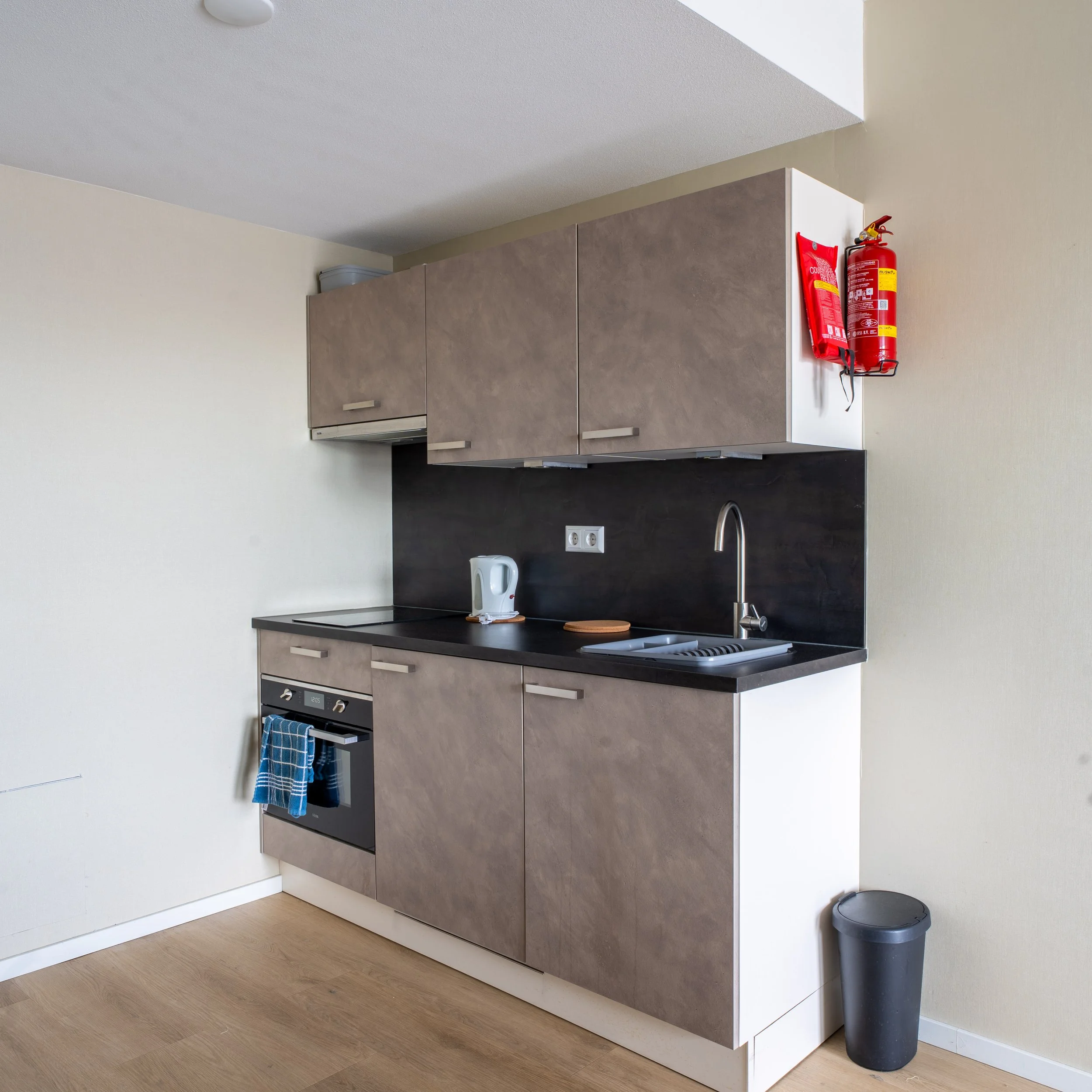 Small kitchen with beige cabinets, black countertop, kettle, dish rack, sink, and a towel hanging on the oven handle. Fire extinguisher mounted on the wall and trash can on the floor.