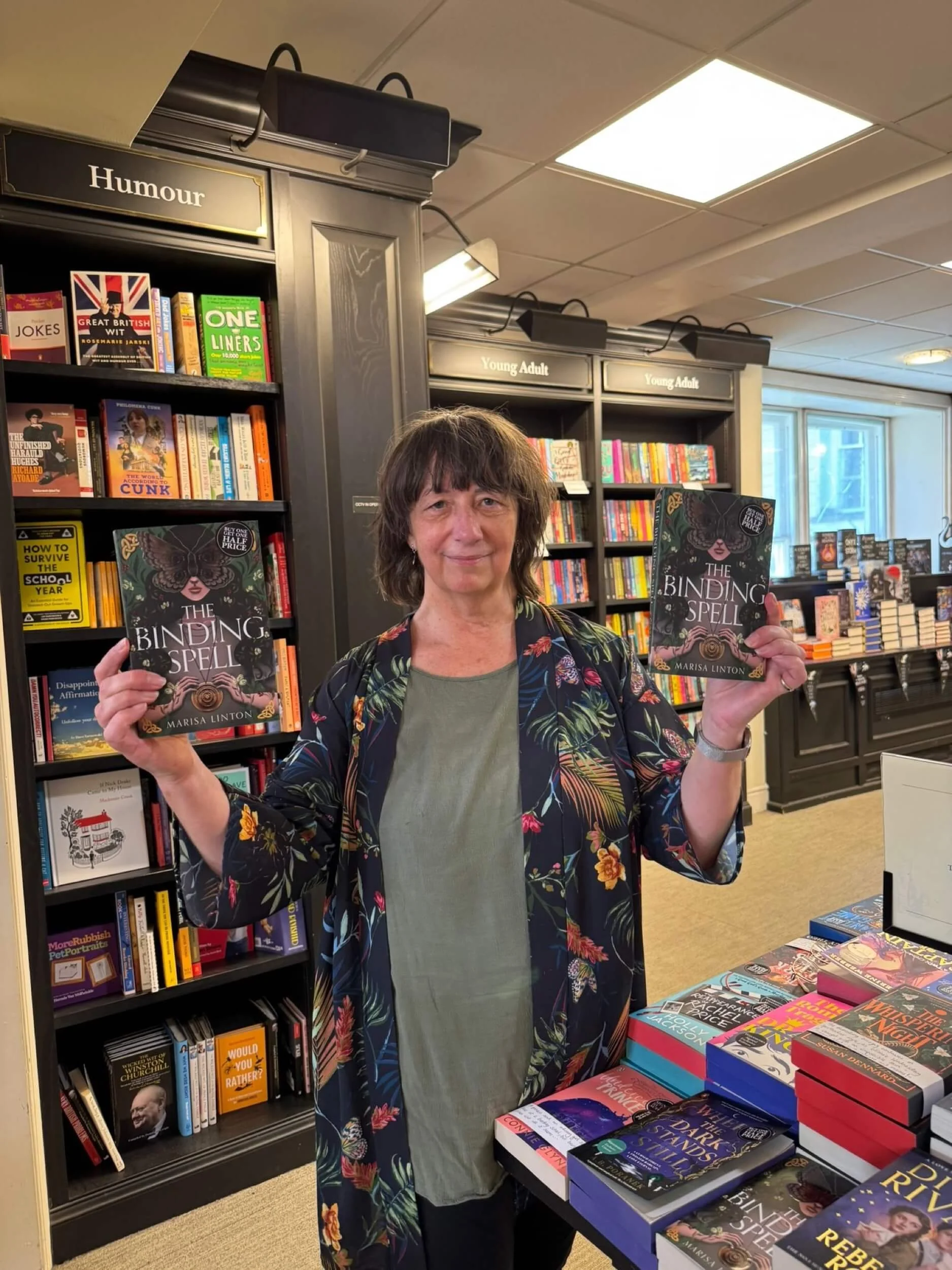 A woman holding two copies of the book titled 'The Binding Spell' in a bookstore. She is standing in front of bookshelves labeled 'Humour' and 'Young Adult' and a table filled with many books.