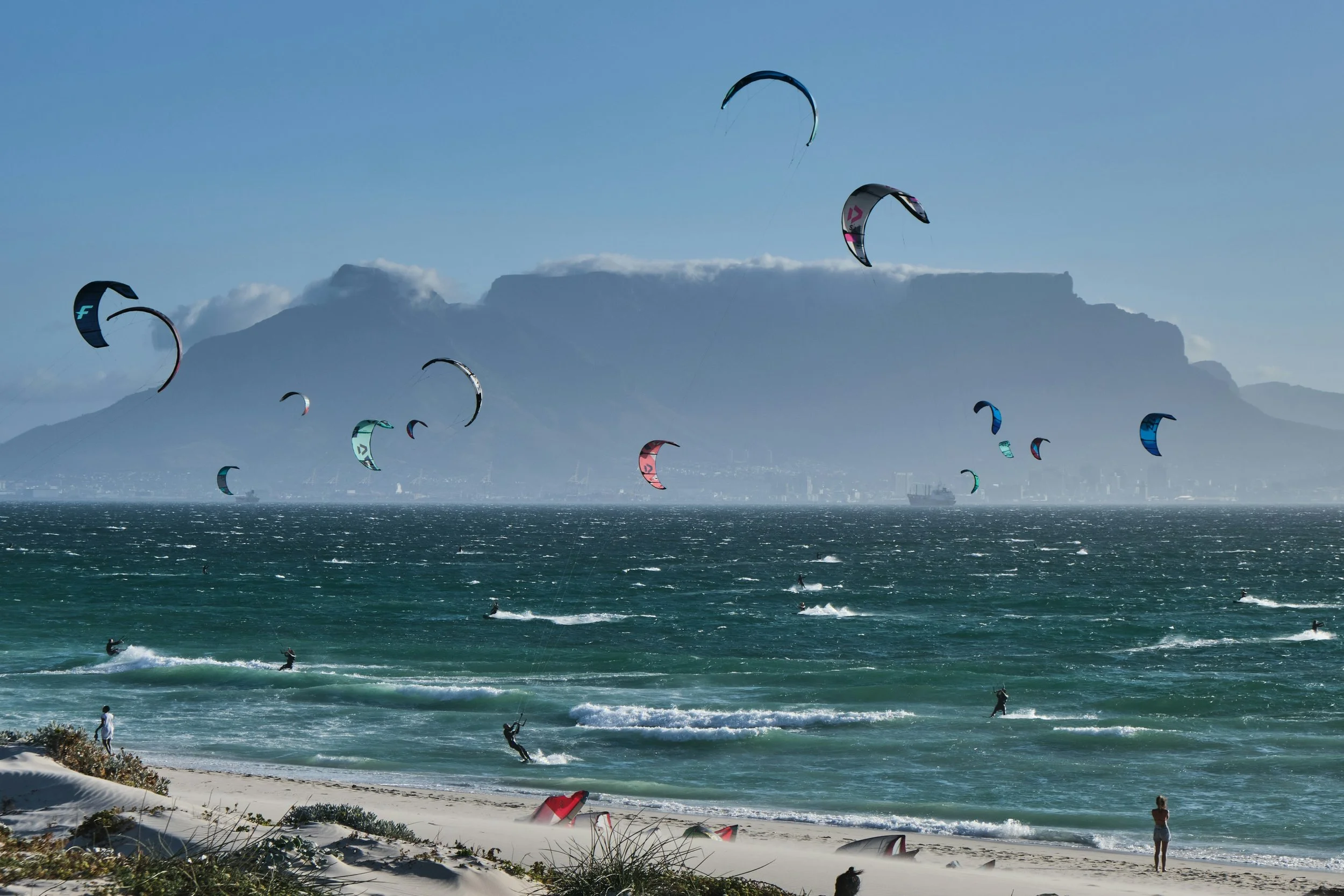 Kite surfers on the ocean near a sandy beach with a mountain in the background, some people watching from the shore.