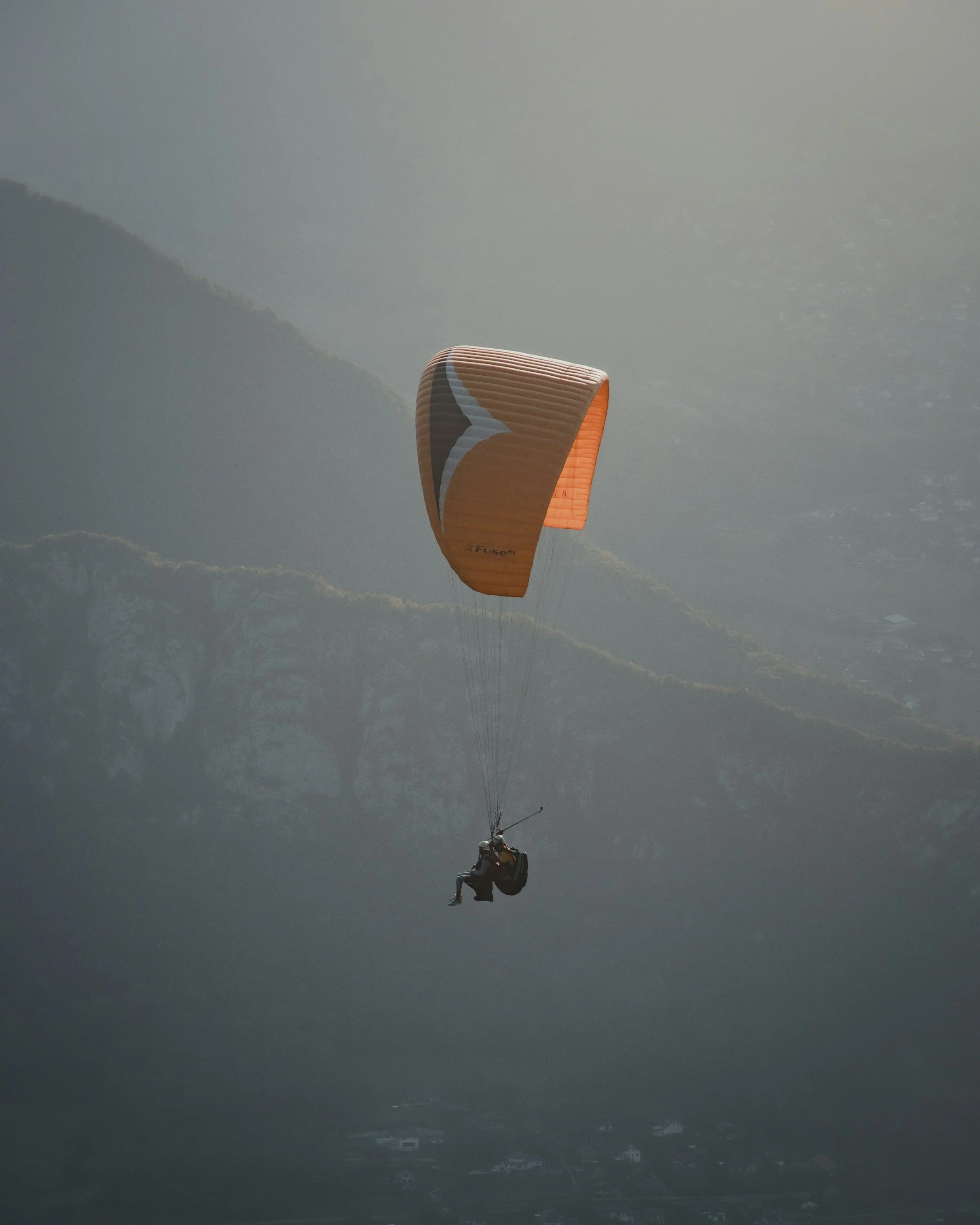 A person paragliding with an orange wing over mountainous terrain at sunset.