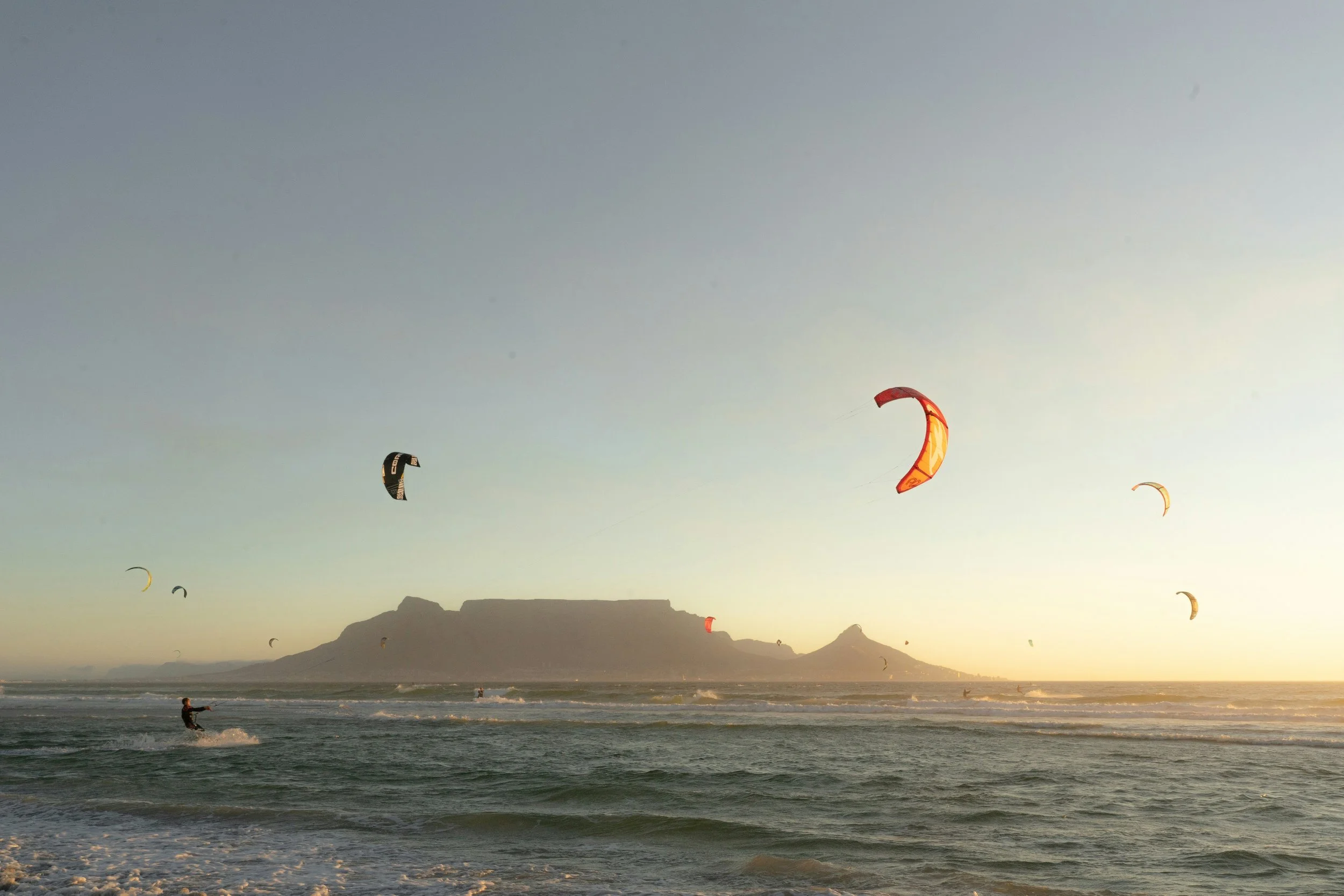 Kite surfers riding the waves at sunset near a mountain in the distance.
