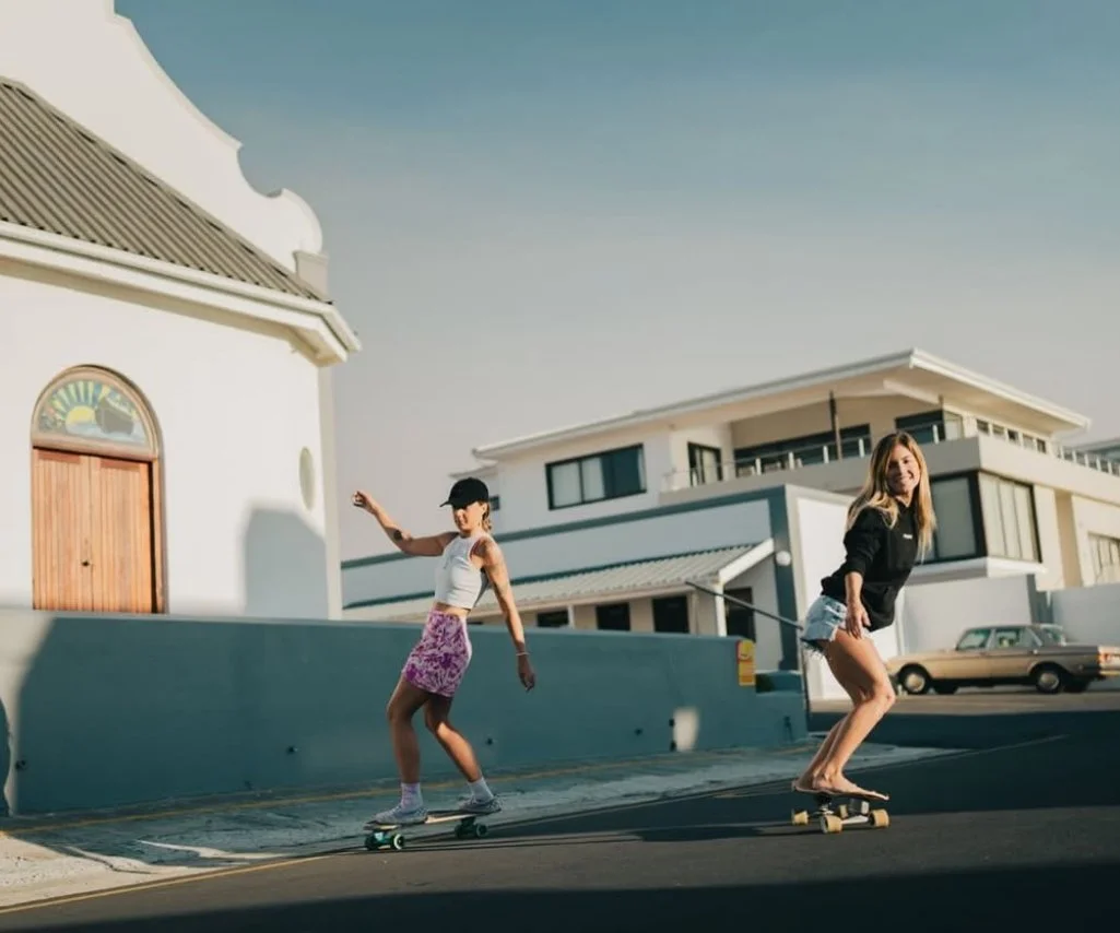 Two young women skateboarding on a street in front of modern white buildings under a clear blue sky.