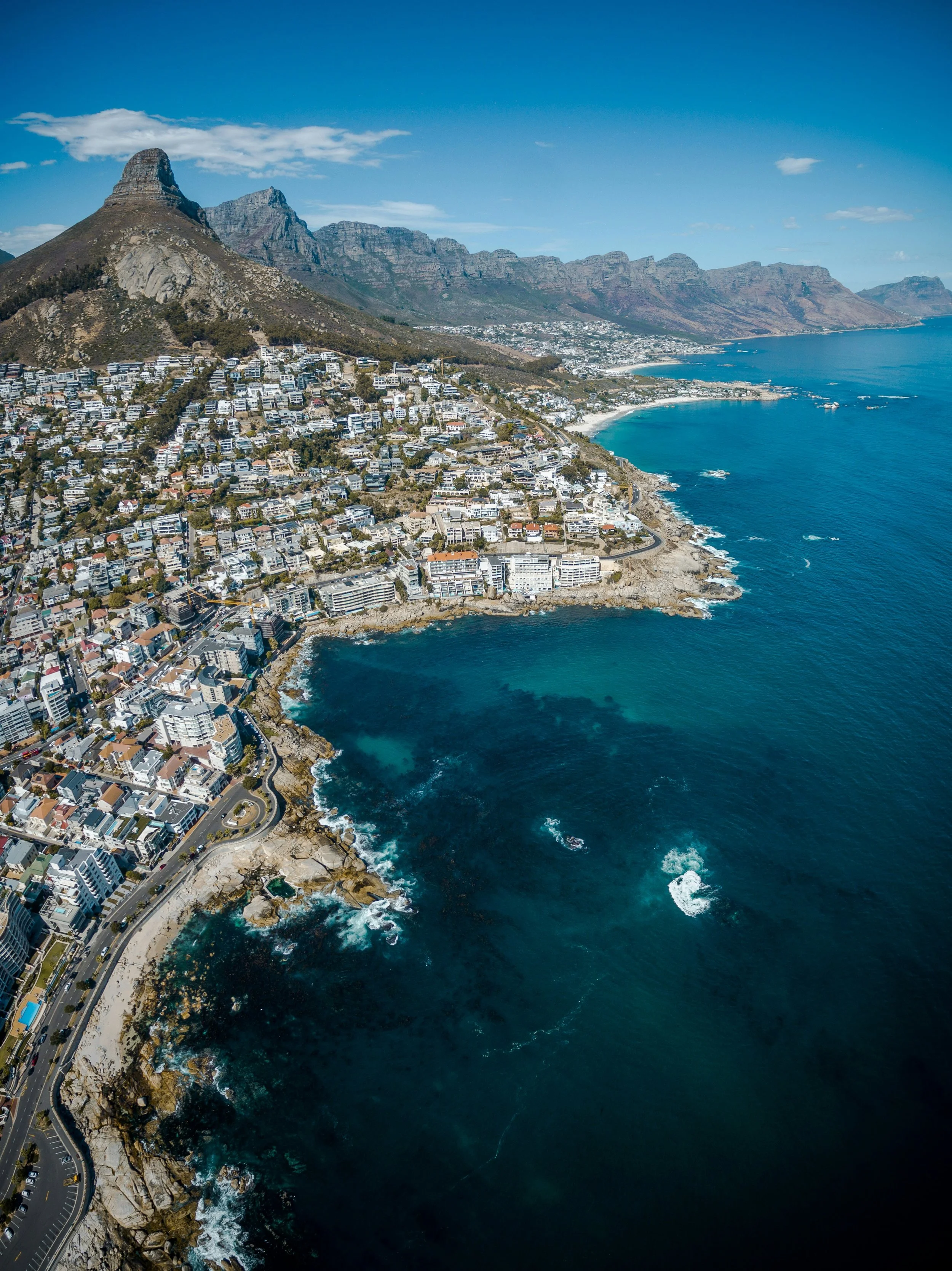 Aerial view of a coastal city with residential buildings on a hillside, rocky shoreline, and blue ocean under a clear sky, with mountains in the background.
