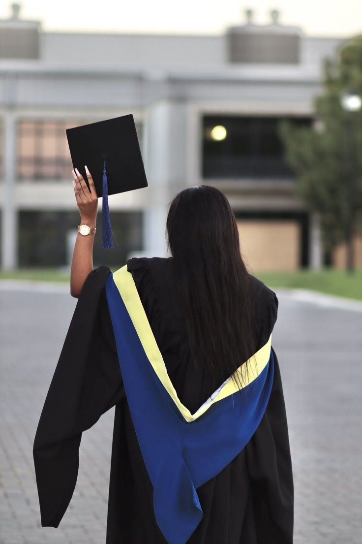 Graduation student in cap and gown holding a mortarboard, standing outdoors with a blurred building in the background.