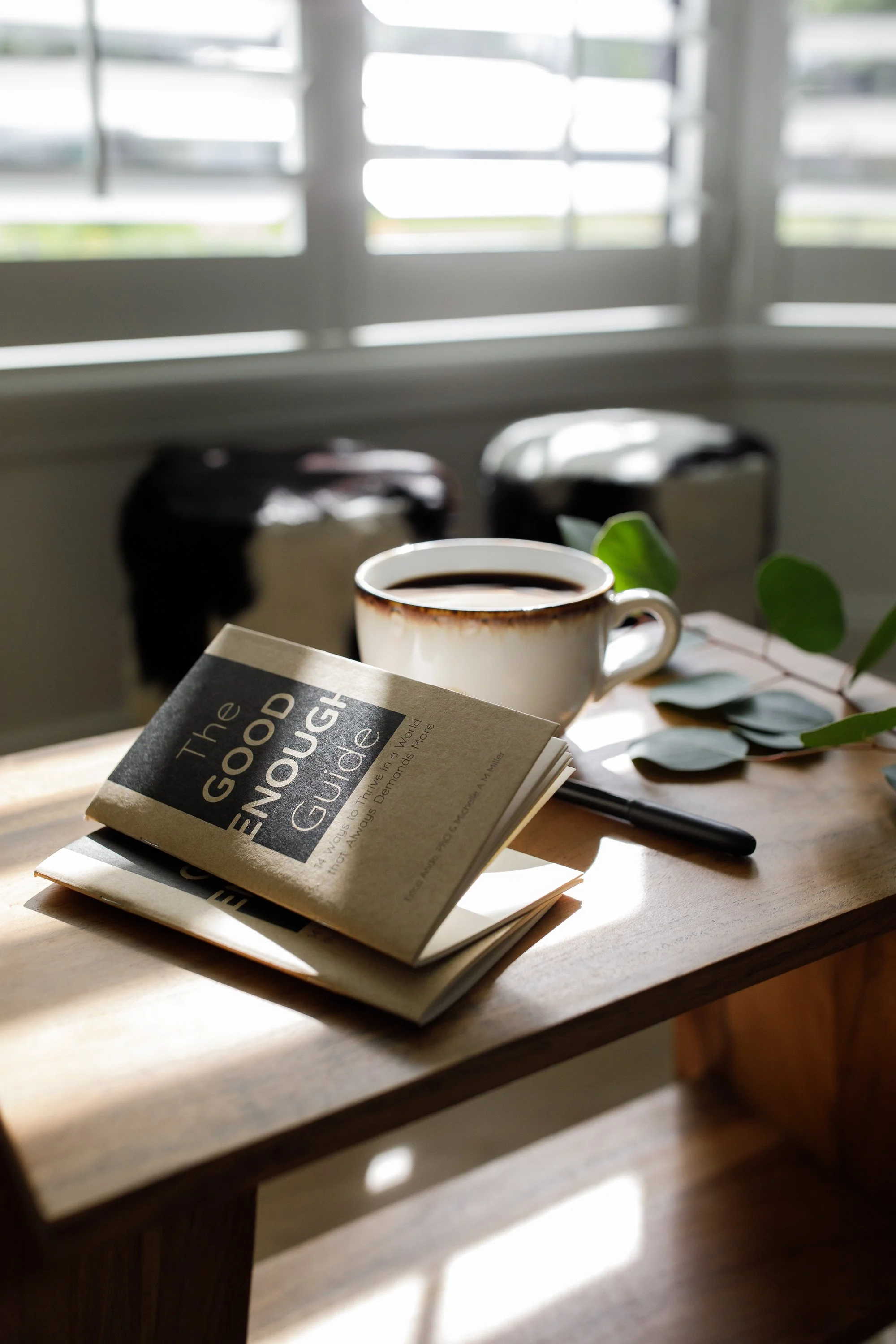 The Good Enough Guide journal, a cup of coffee and a pen on a wooden table by a sunlit window