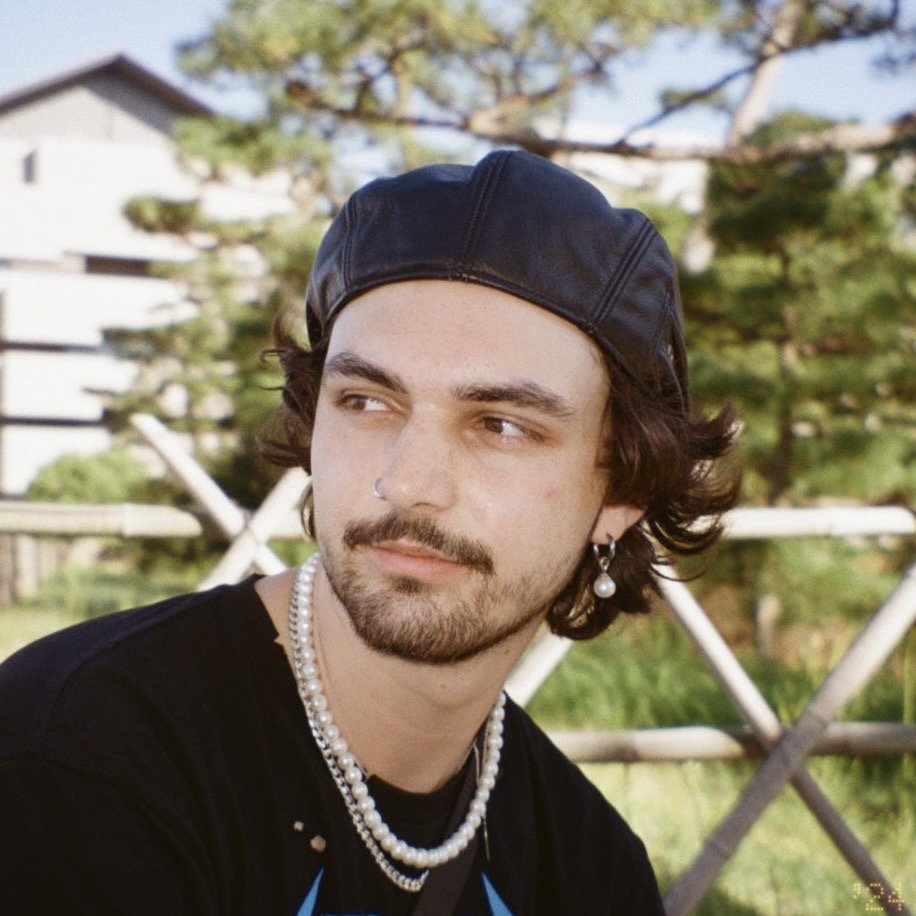 A young man with dark hair and a beard looking to the side, wearing a black leather cap, pearl necklace, and earrings, outdoors with trees and a white building in the background.