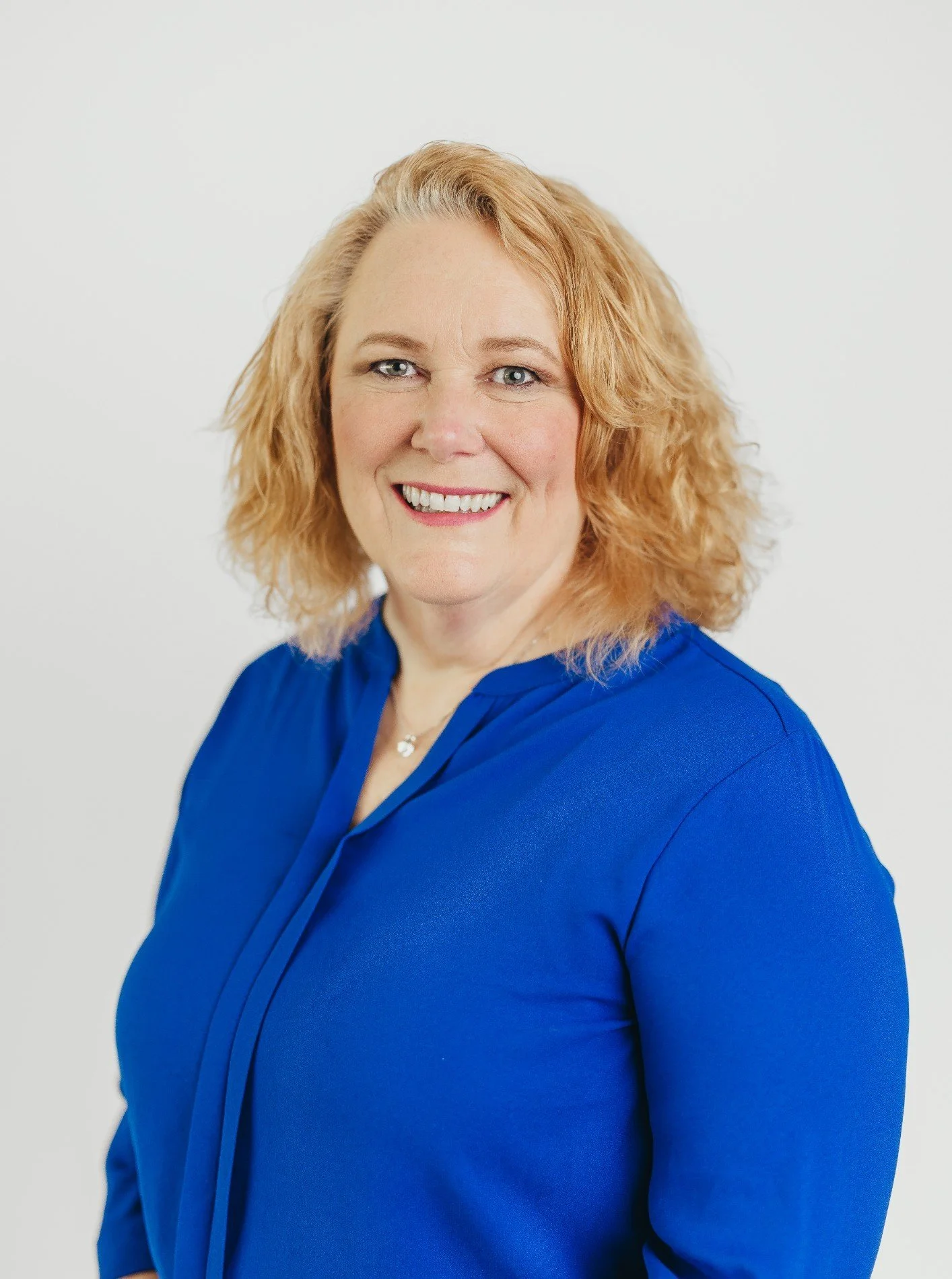 A smiling woman with shoulder-length curly blonde hair, wearing a royal blue blouse, standing against a plain white background.