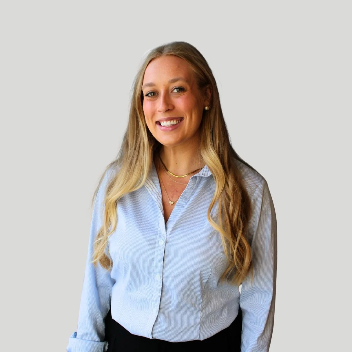 A woman with long blonde hair wearing a light blue button-up shirt and gold jewelry, smiling against a plain light gray background.