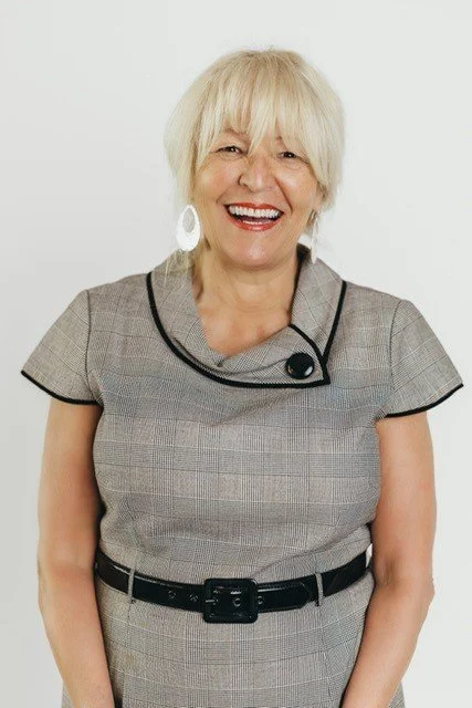 A smiling senior woman with short white hair, wearing a gray plaid dress with black trim and a black belt, against a white background.