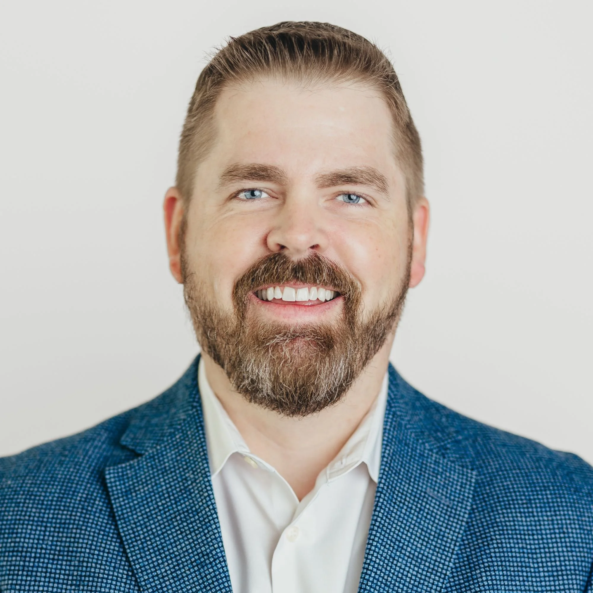 A smiling man with a beard and mustache, blue eyes, wearing a white shirt and blue blazer, against a plain light background.