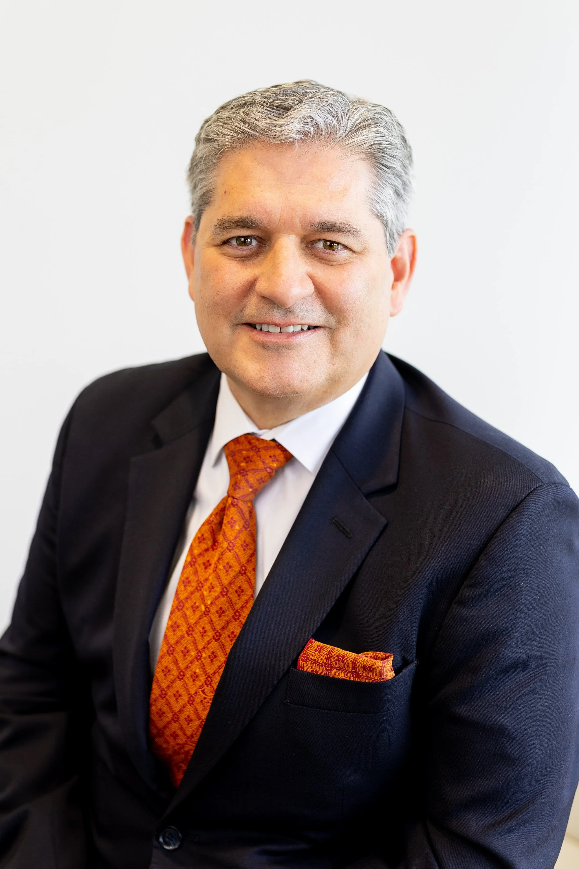 A middle-aged man with gray hair, wearing a dark suit, white shirt, and an orange patterned tie and pocket square, smiling in a professional portrait.