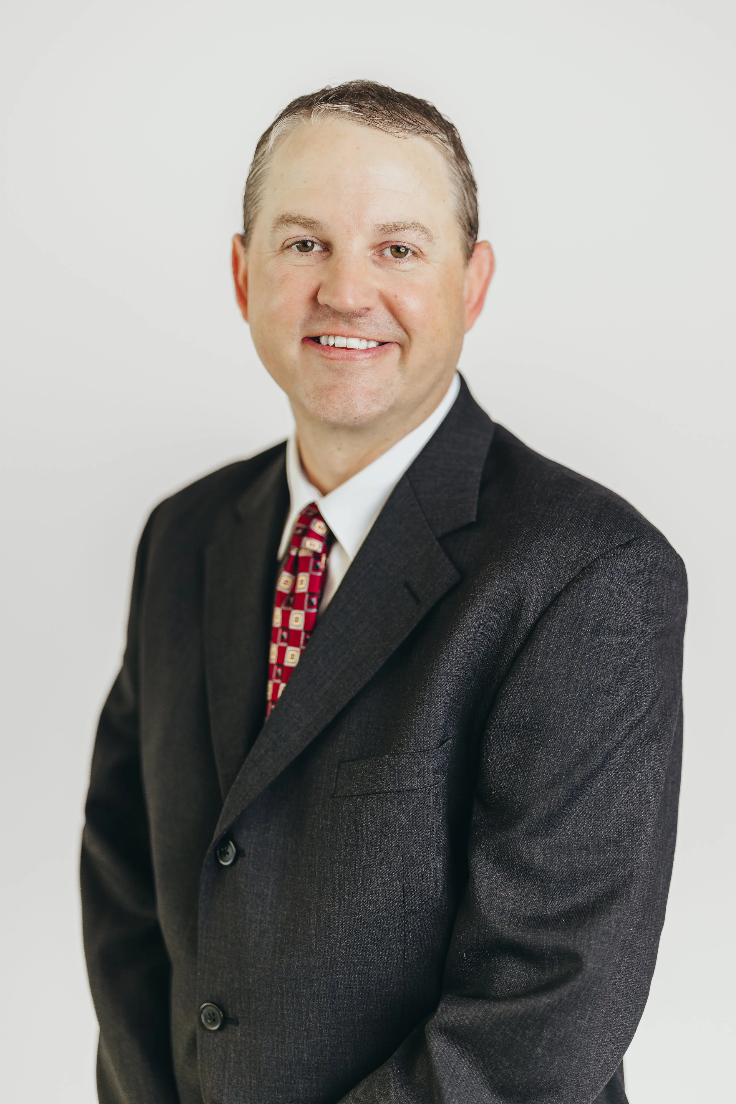 Professional headshot of a man in a dark suit, white shirt, and colorful patterned tie, smiling against a plain white background.