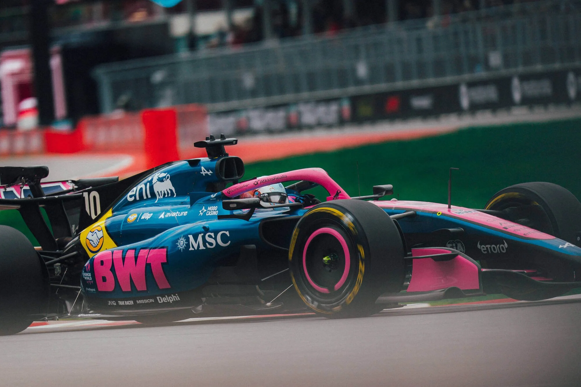 A Formula 1 race car with a blue, pink, and black livery on a race track. The car features prominent sponsor logos including BWT, MSC, and others. The driver is wearing a helmet and is visible through the cockpit. The background shows a blurred grandstand and barriers.