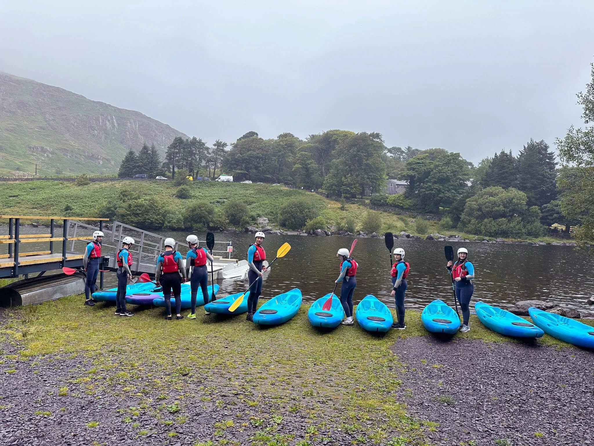 A group of people wearing life jackets and helmets standing next to blue kayaks by a river, with trees, hills, and a cloudy sky in the background.