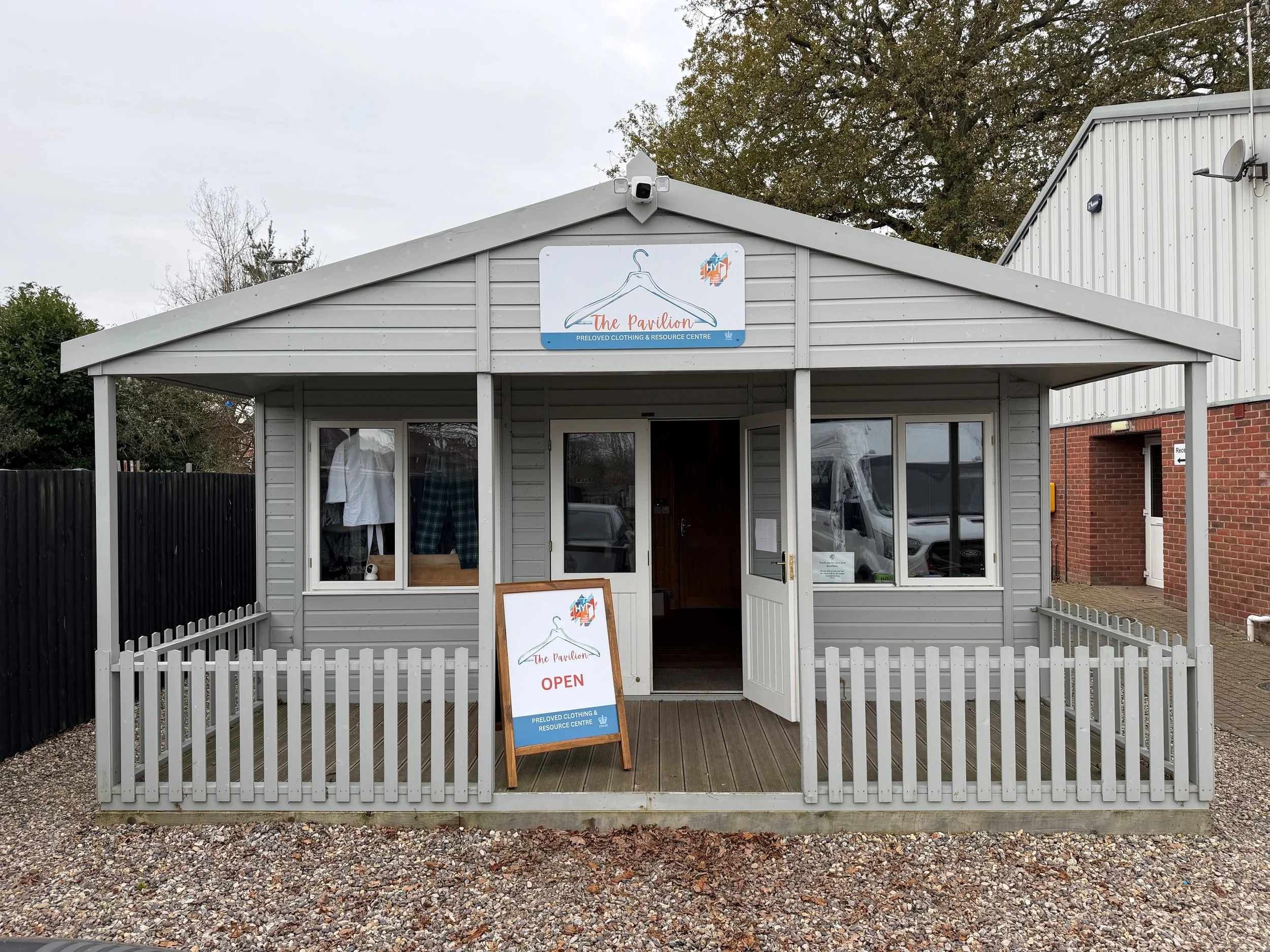 Small building with a white picket fence, sign on top reading 'The Pavilion', open sign on a stand outside, and a window display of clothing.