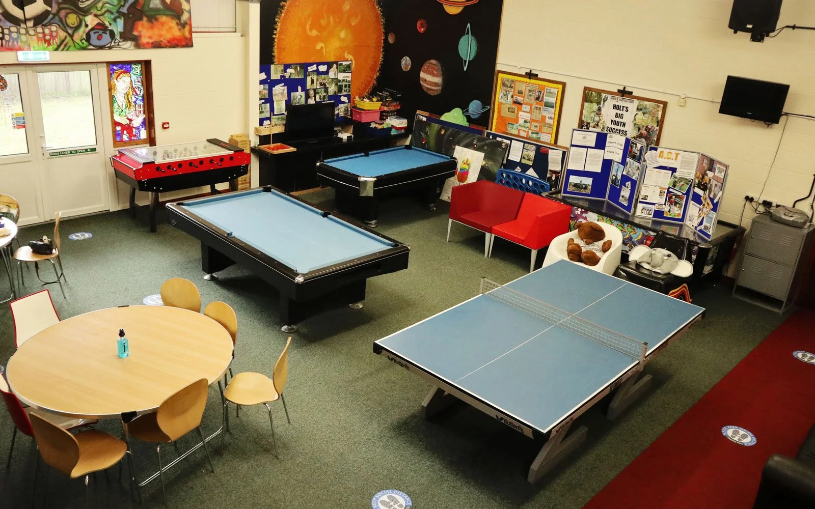 Room with recreational activities, including two pool tables, a table tennis table, a foosball table, a red and white seating area, a round table with chairs, and educational display boards on the walls.