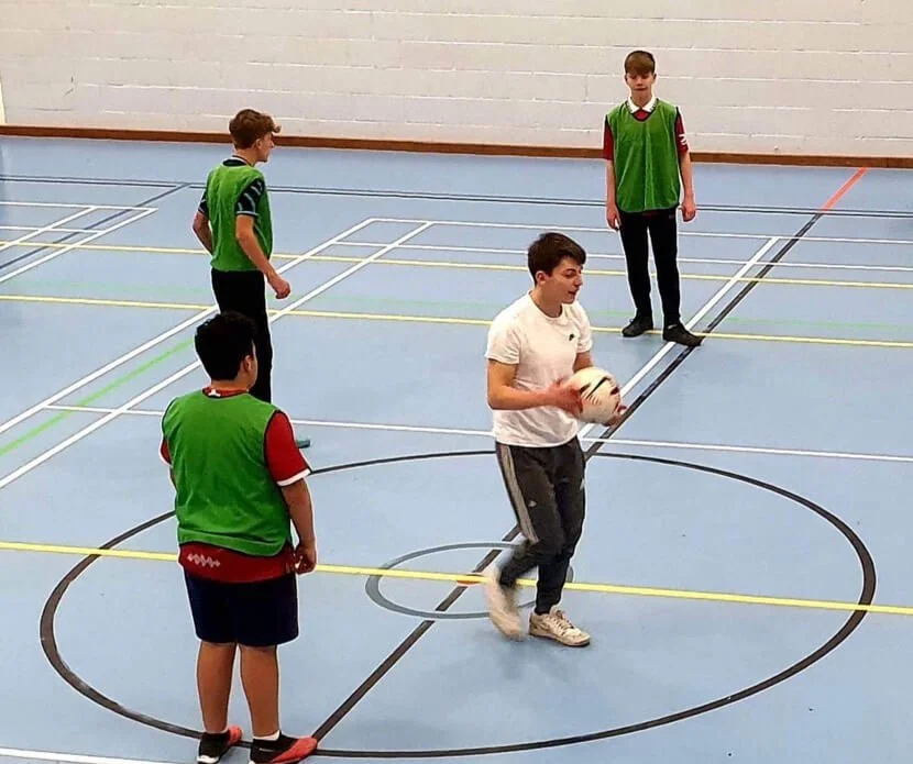 Four boys in sports attire on an indoor court, with one holding a volleyball, possibly preparing for a game or practice.