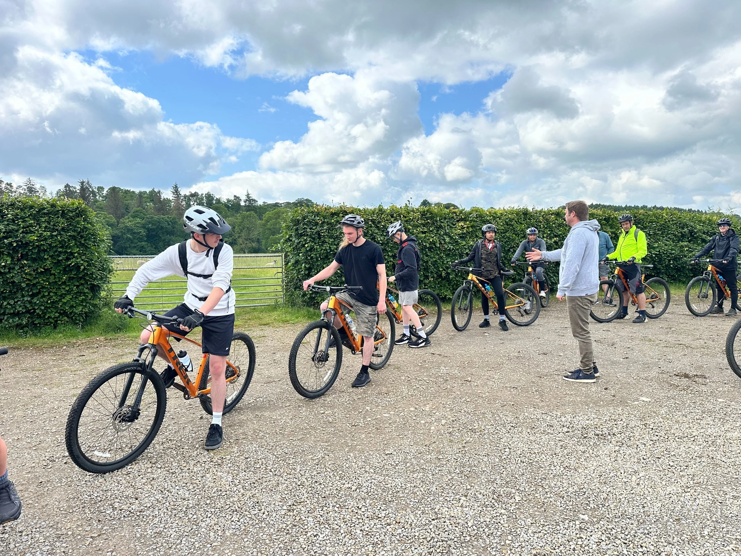 A group of people with bikes gathering outdoors, some listening to a person in a gray hoodie talking, in a setting with a gravel ground, green hedges, and a cloudy sky.