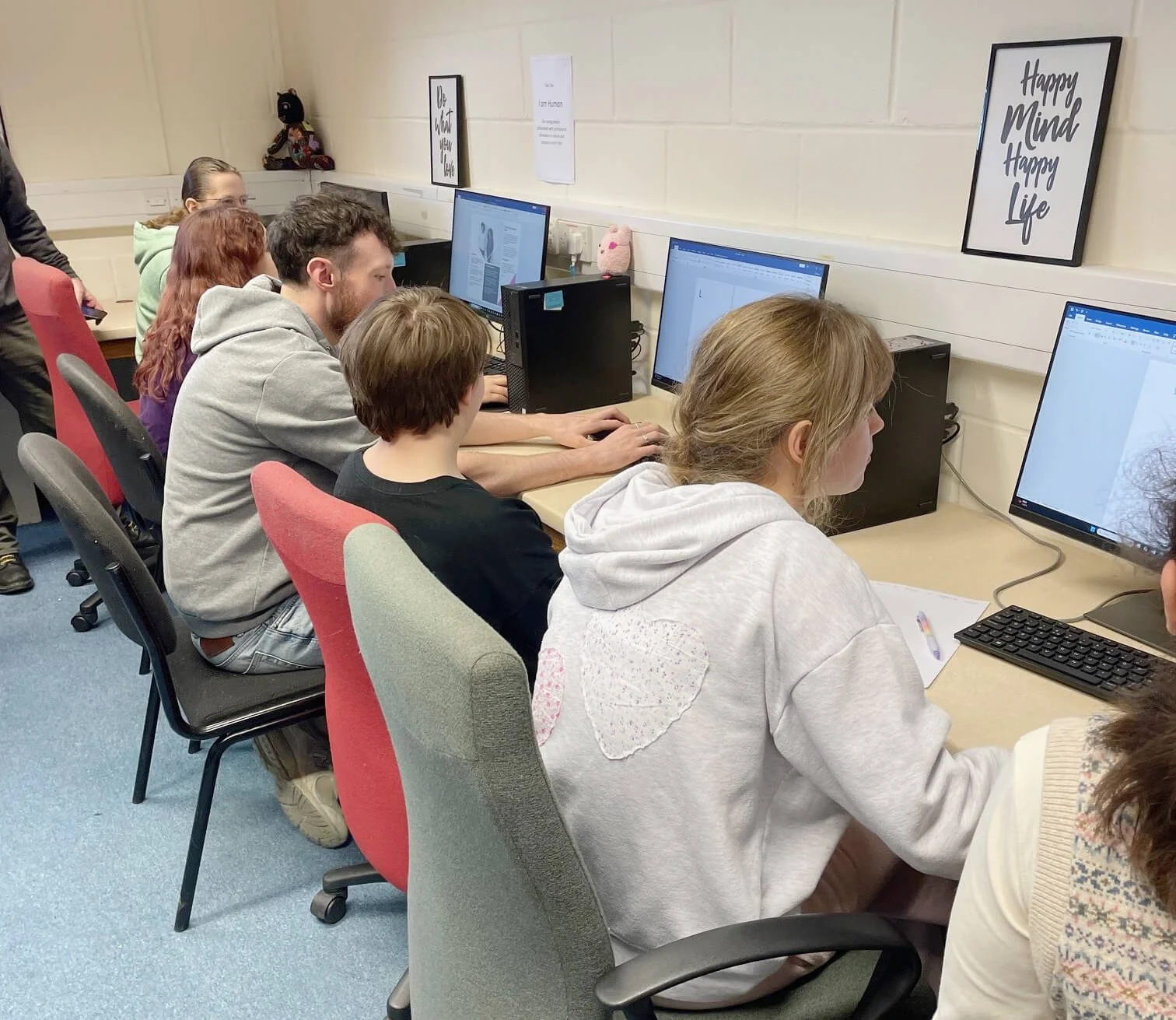 A group of six people working on computers in a classroom, with framed quotes on the wall reading 'Be What You Can Be' and 'Happy Mind Happy Life'.