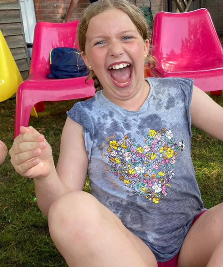 A young girl with blonde hair and blue eyes laughing outdoors, sitting on grass with pink chairs in the background