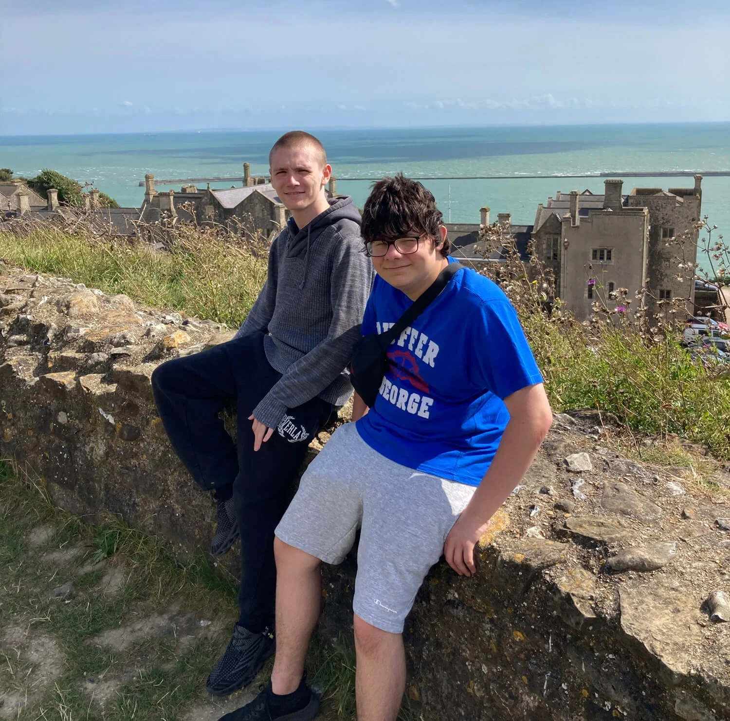 Two boys sitting on a stone wall with a coastal town and ocean in the background.