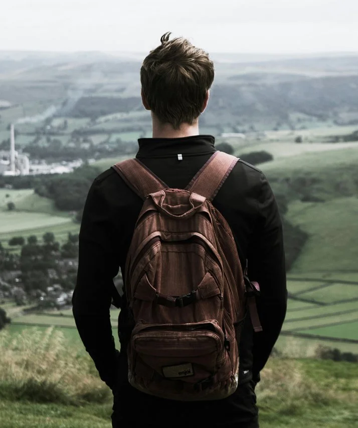 A person with short hair wearing a black jacket and a brown backpack standing on a hilltop, overlooking a vast green landscape with fields and hills.