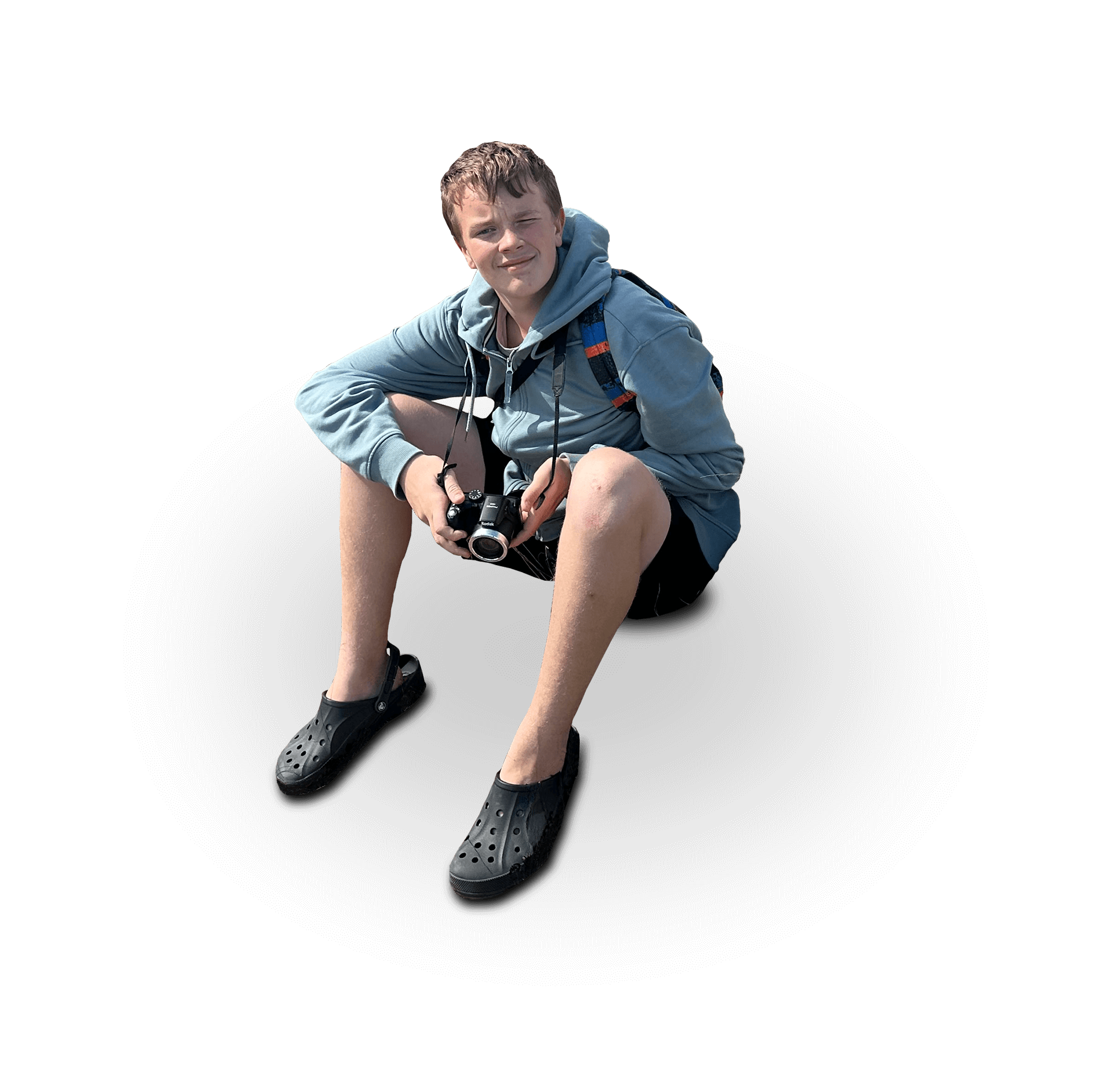A young man sitting against a black background, wearing a blue hoodie, shorts, and black Crocs, holding a camera in his hands and looking at the camera with a slight smile.