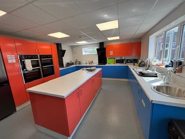 Colorful kitchen with red and blue cabinets, white countertops, and stainless steel sink, with windows on the side and various appliances on the counters.
