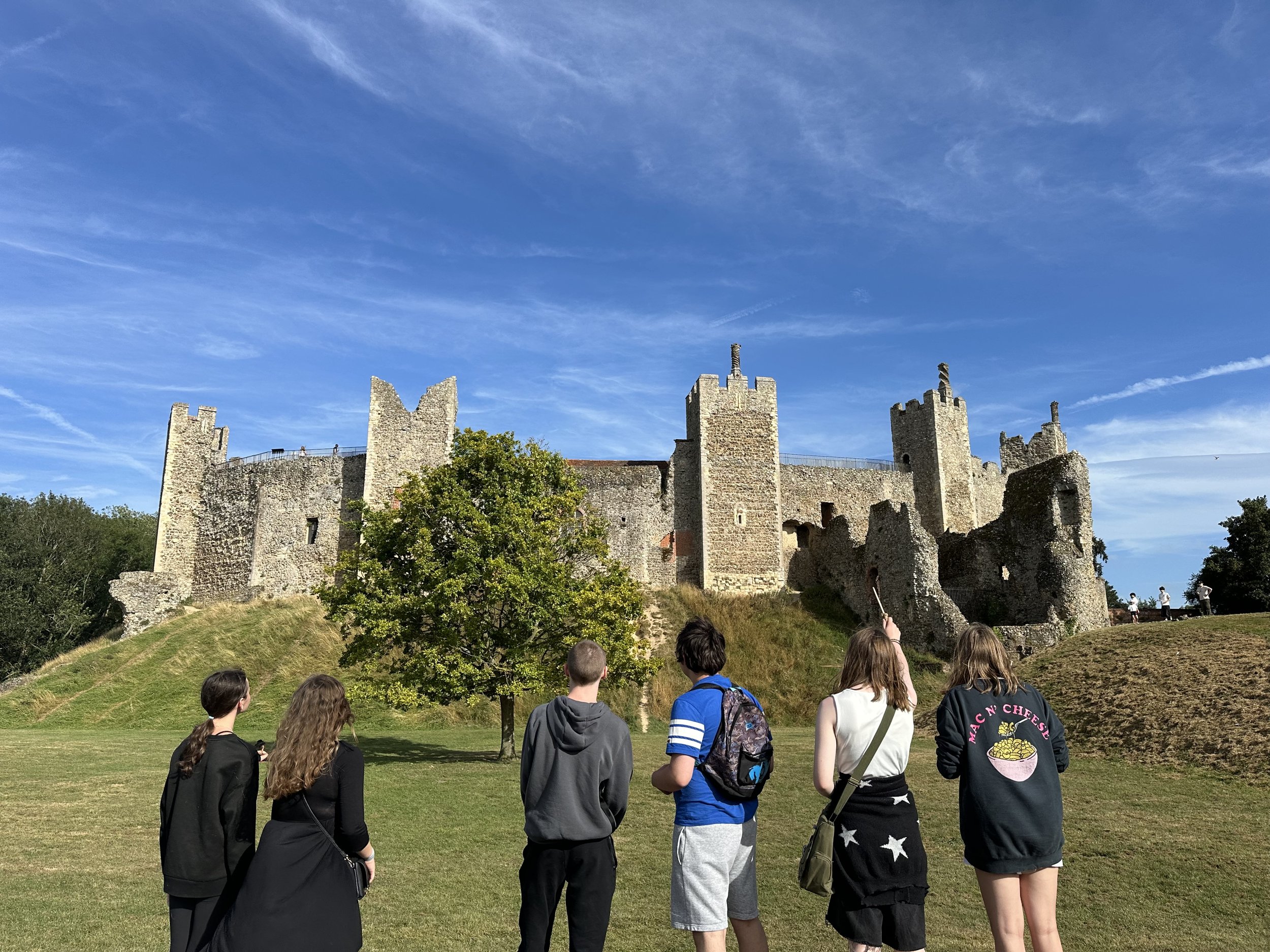 A group of seven tourists, including three girls and four boys, stand on a grassy hill in front of an ancient castle with stone walls and towers under a blue sky with clouds.