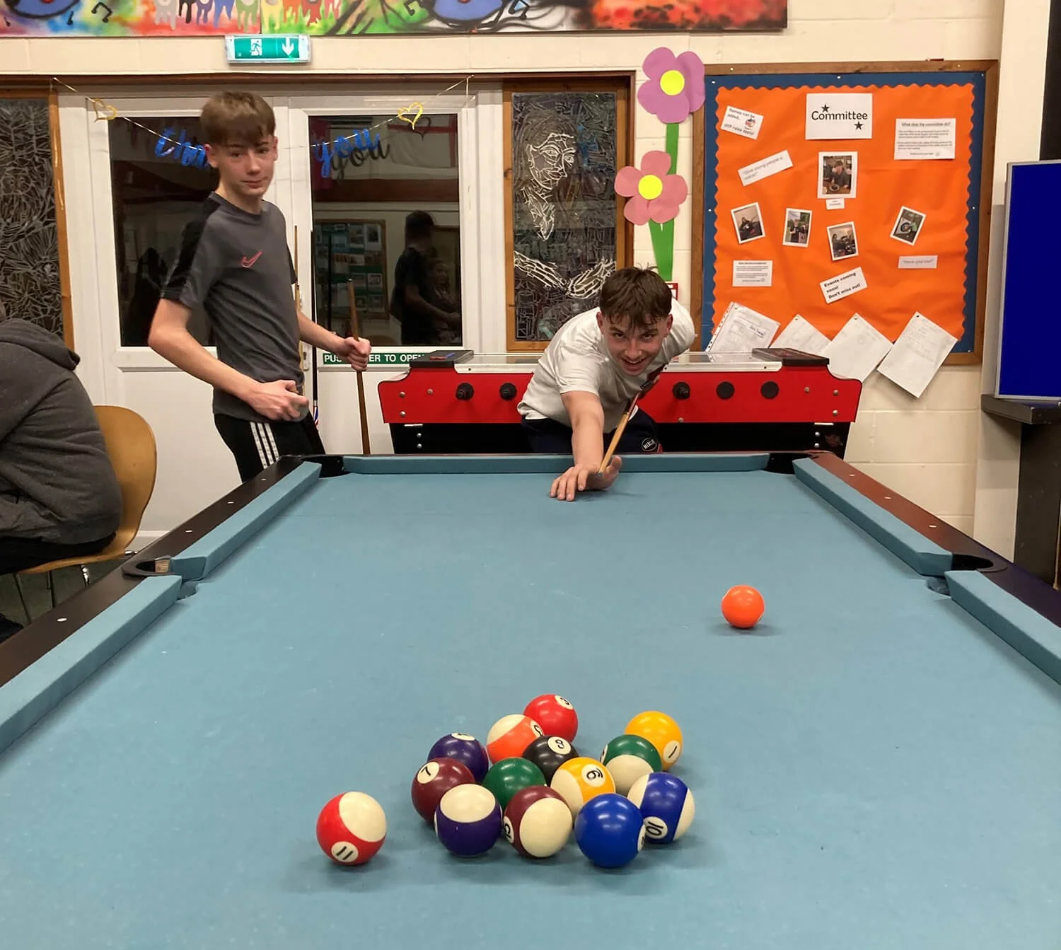 Two boys playing pool in a recreational room with colorful decorations and a bulletin board in the background. One boy is aiming a shot at the cue ball, while the other stands nearby. The pool table has a set of balls arranged at one end.