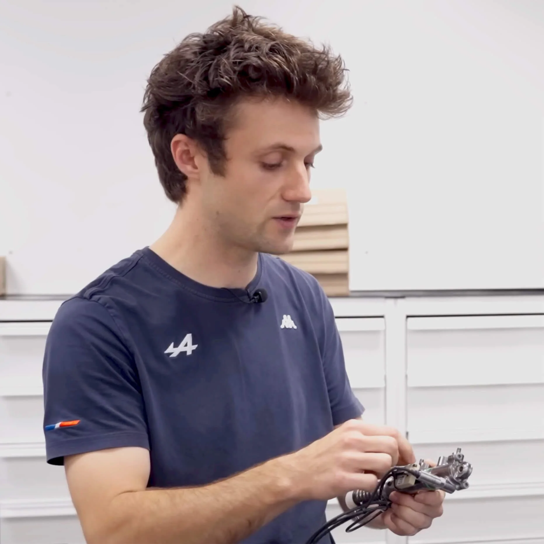 A young man with tousled brown hair looking down at an object he is holding, wearing a navy blue T-shirt with white logos, standing in front of white drawers or cabinets in a room with a white wall.