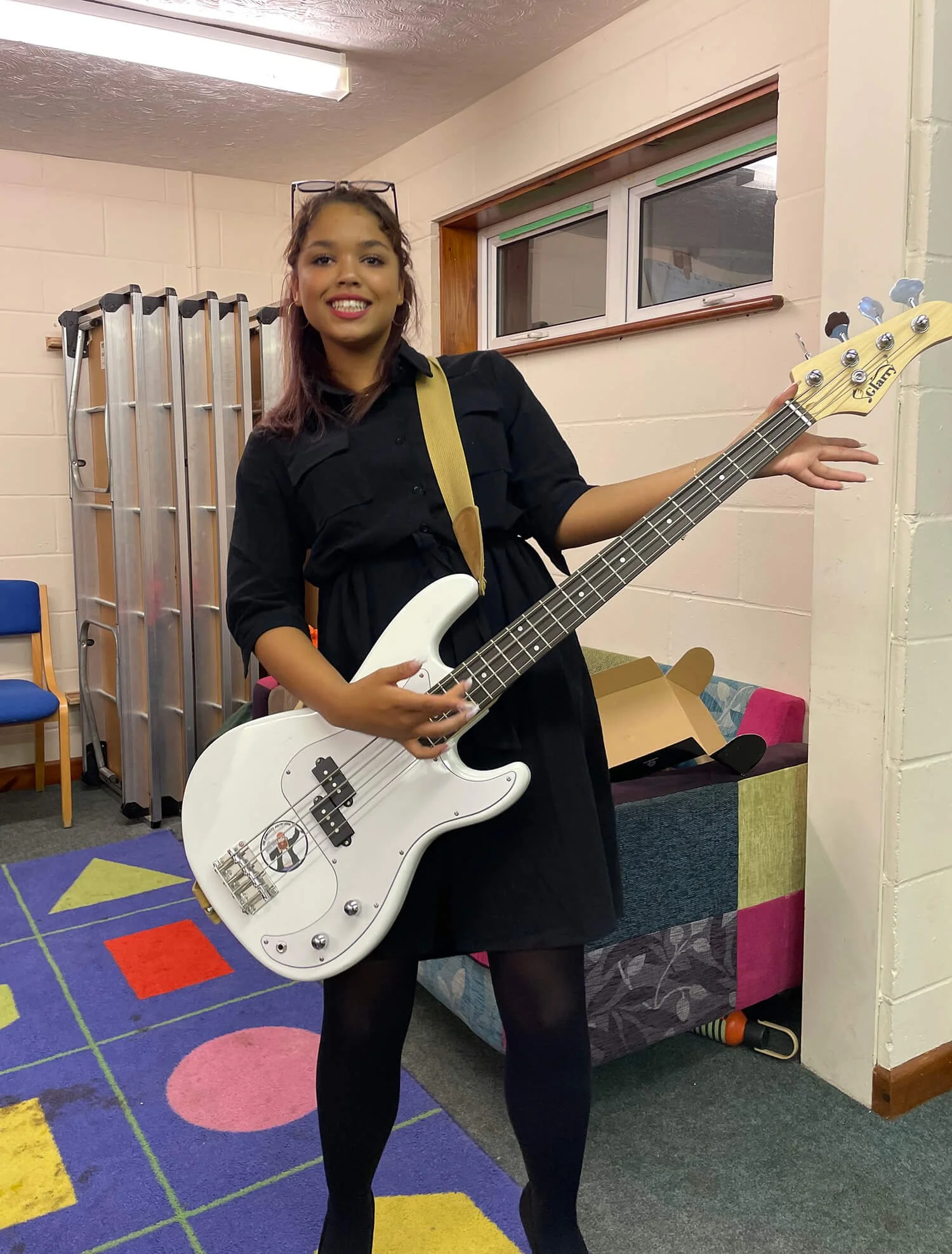 A young girl with glasses on her head holding a white bass guitar in a room with colorful carpet, chairs, and a cardboard box behind her.