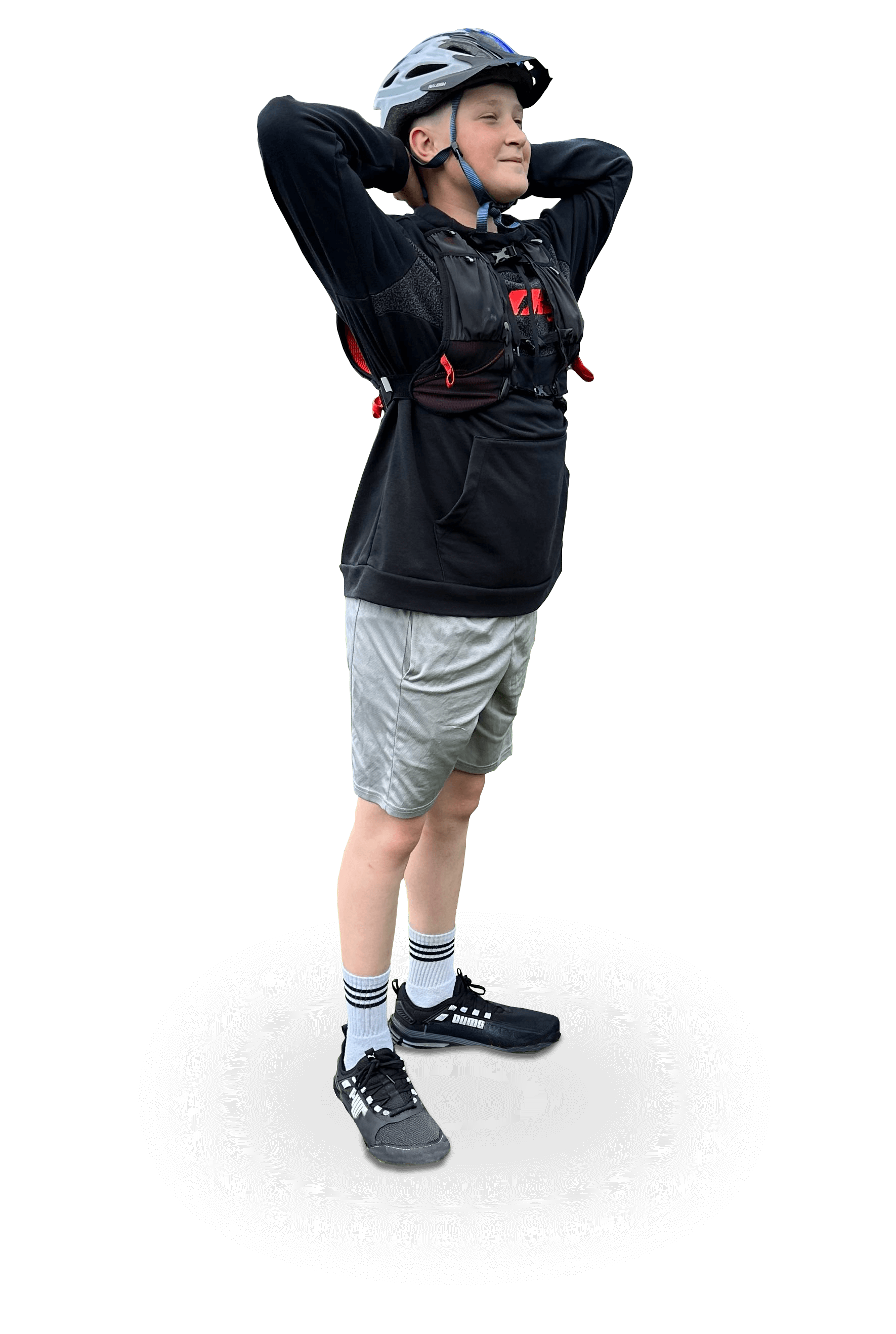 A young boy standing with hands behind his head, wearing a bicycle helmet, black hoodie, gray shorts, white socks with black stripes, and black sneakers, against a black background.