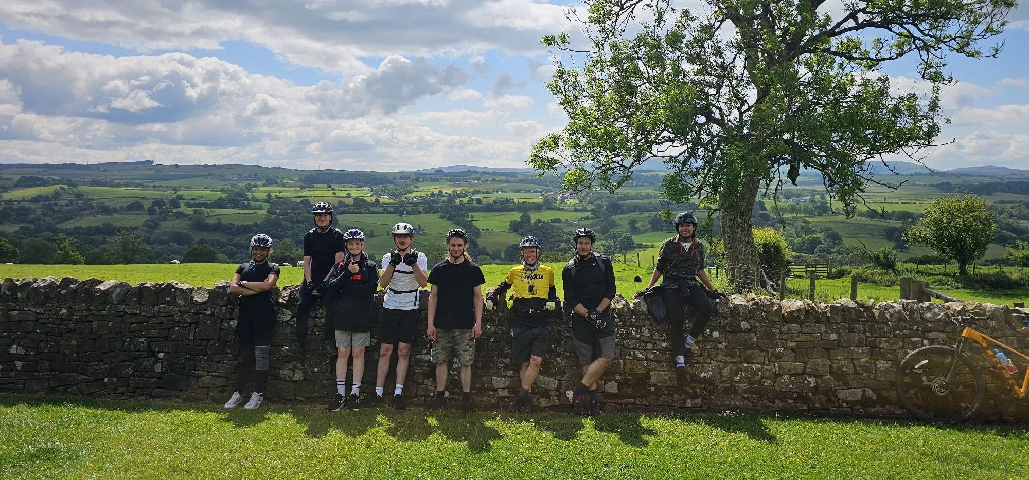A group of eight cyclists wearing helmets and casual clothes, standing and sitting near a stone wall in a rural landscape with green fields, trees, and rolling hills under a partly cloudy sky. A yellow bicycle is partially visible on the right side of the image.
