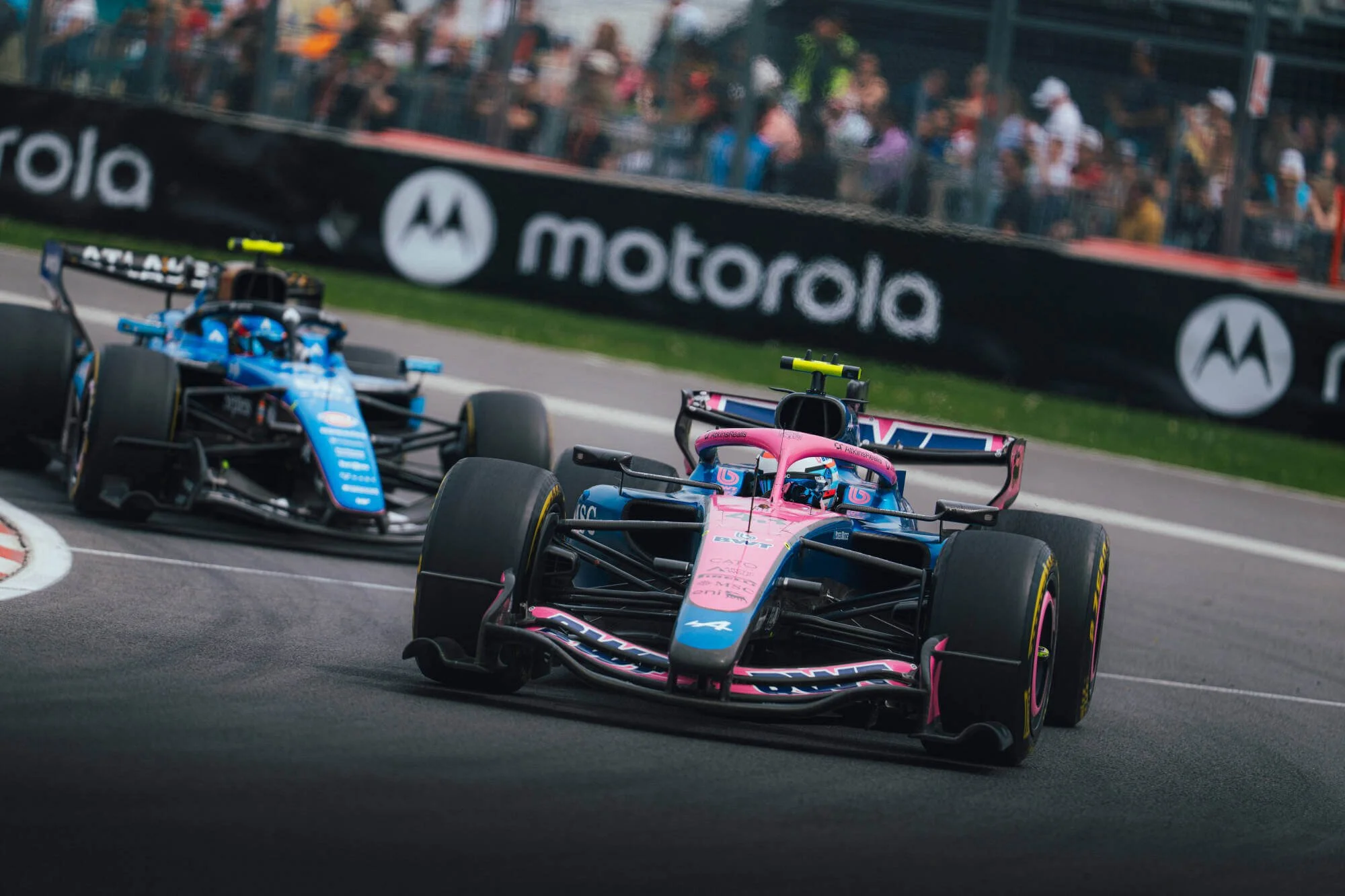 Two Formula 2 race cars, one in pink and blue and the other in blue, racing on a street circuit with a crowd of spectators in the background and large Motorola advertisements along the fence.