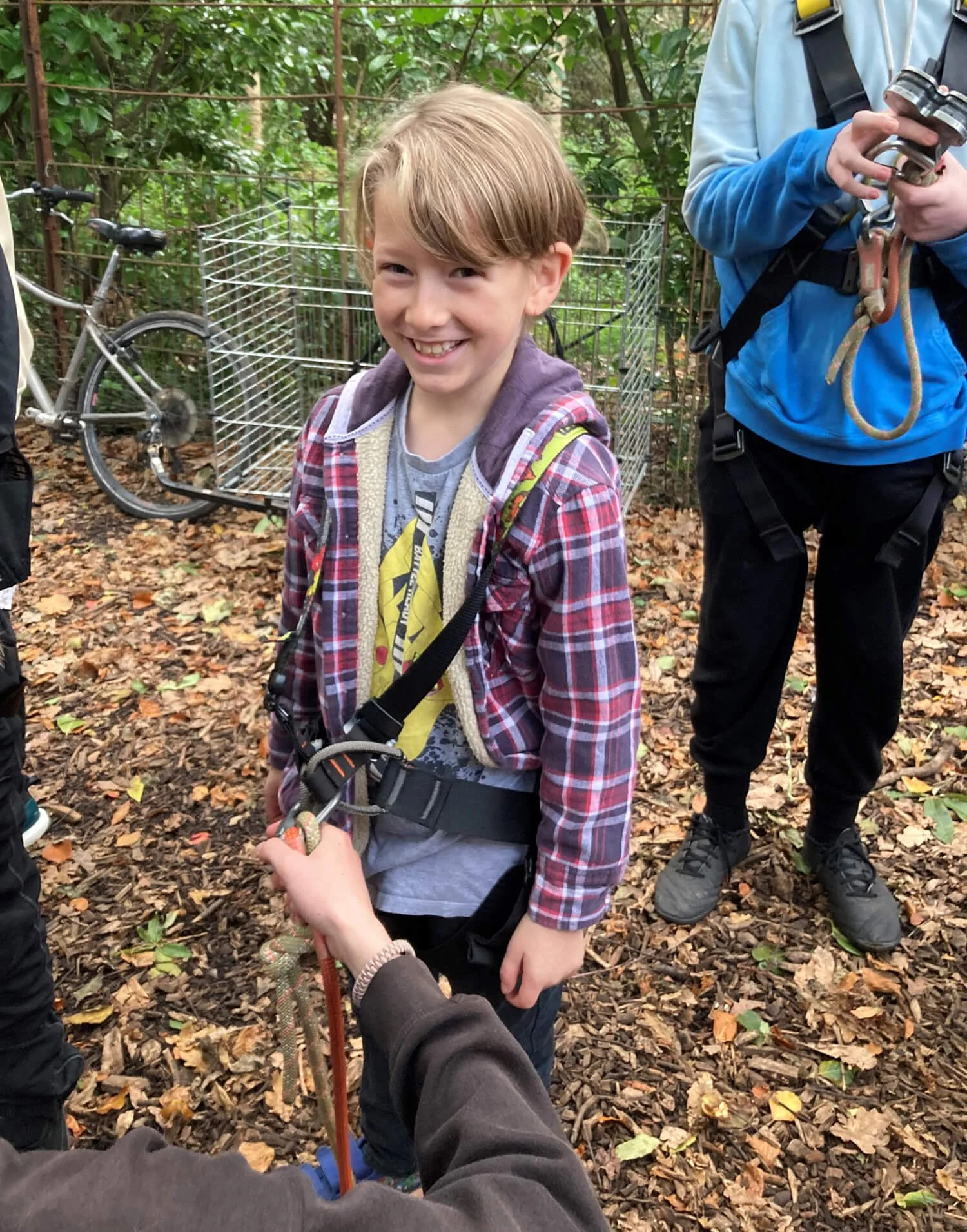 A young boy smiling outdoors in a wooded area, wearing casual clothes and a harness, with a person holding onto a rope.