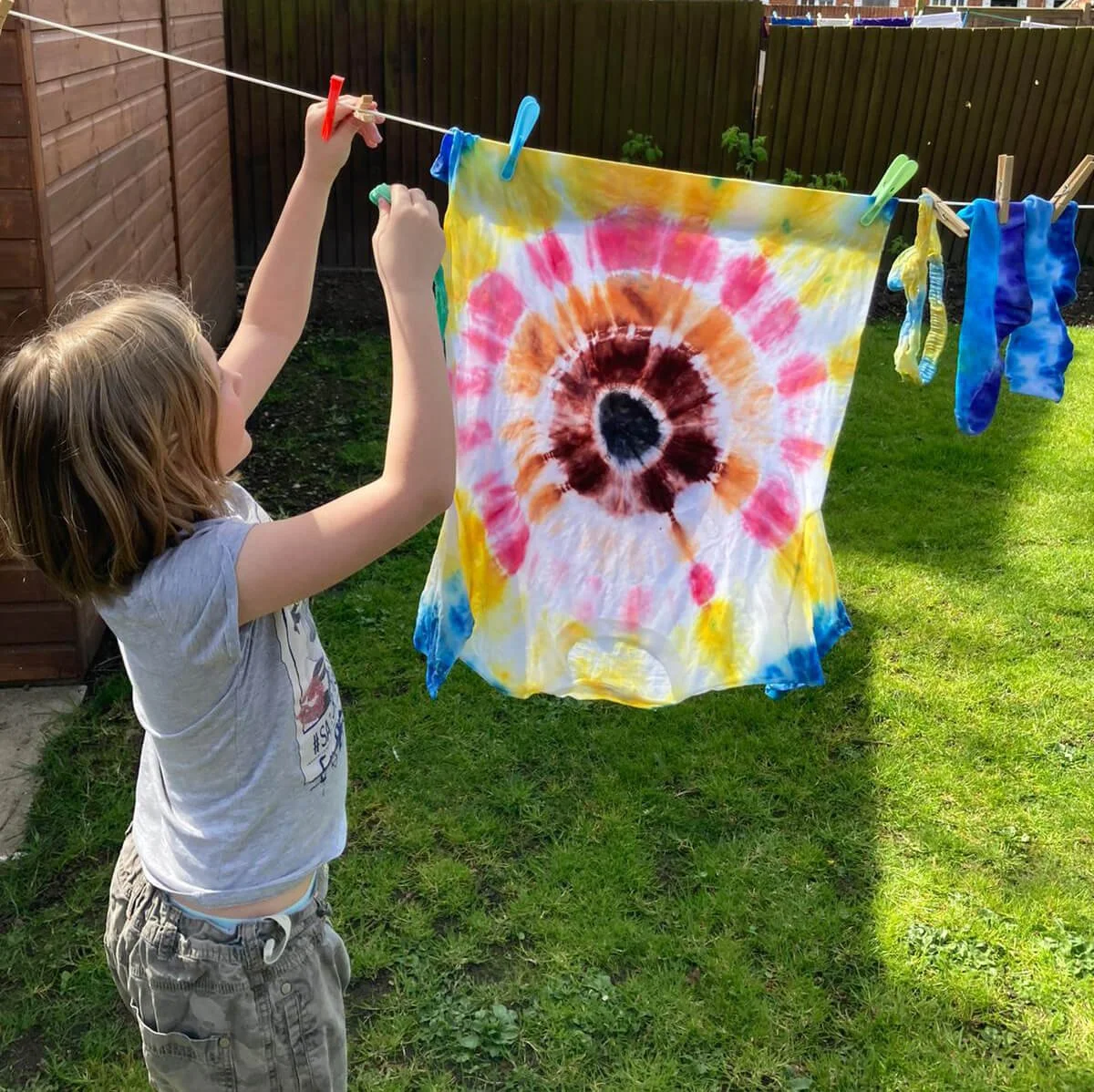 A young girl hanging a tie-dye cloth on a clothesline in her backyard. The cloth features a colorful spiral pattern with pink, orange, brown, and white colors. There are other pieces of clothing or fabric on the line to the right.