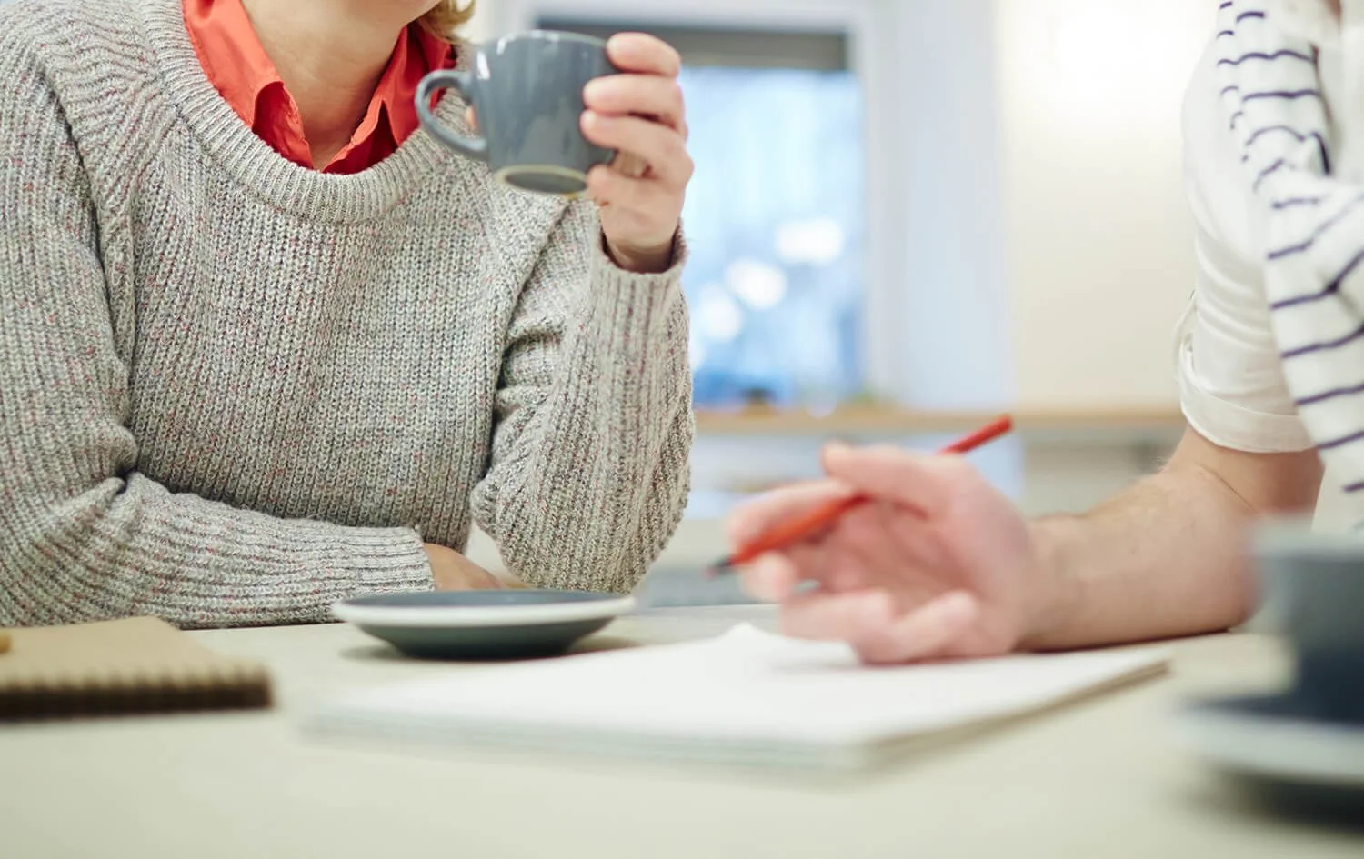 A woman wearing a beige sweater and red shirt drinking coffee while sitting at a table with a man who is writing on paper with a red pen.