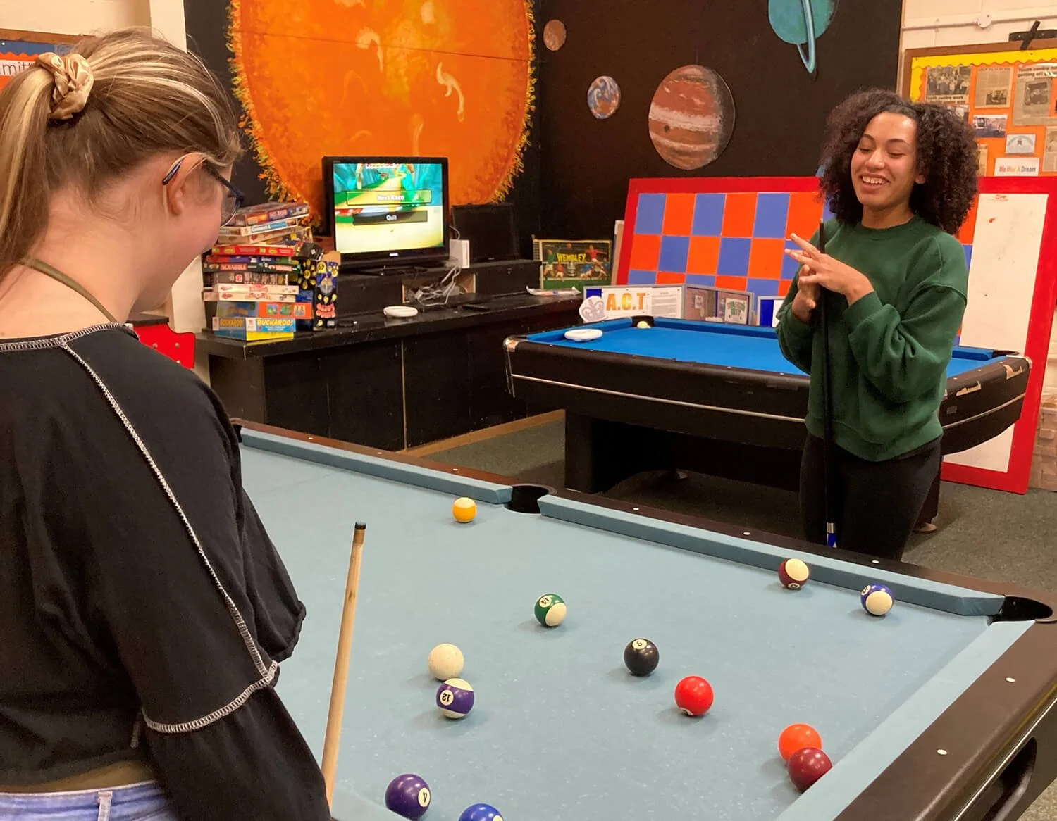 Two girls playing pool in a room filled with books, board games, and space-themed decorations, including a large sun painting and planets on the wall.