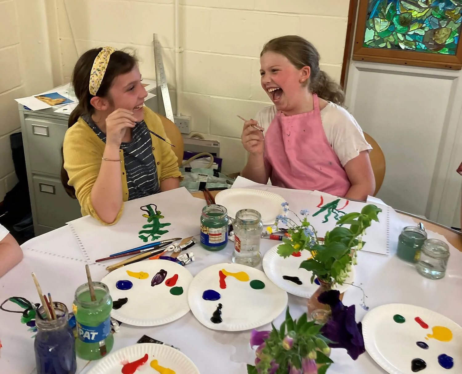 Two girls sitting at a table, laughing and painting on paper plates. The table has paint jars, brushes, and paper plates with colorful paint patterns. There are flowers in vases on the table.