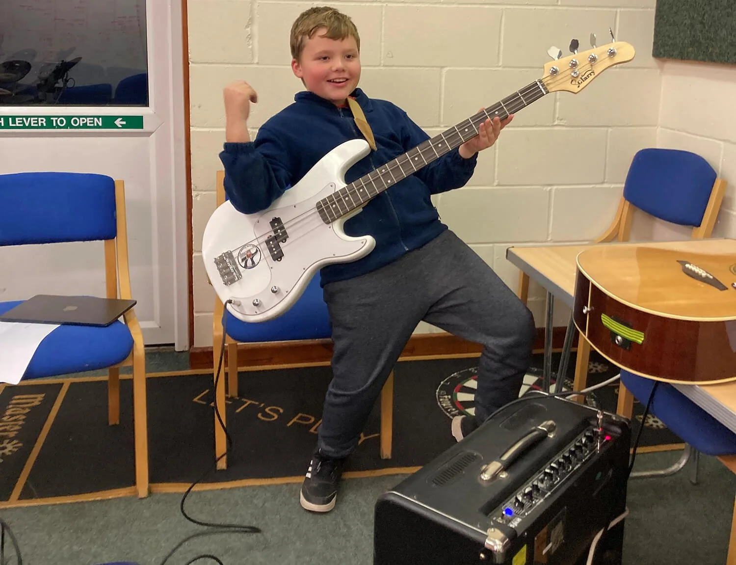 A boy with brown hair wearing a dark blue hoodie, gray sweatpants, and black sneakers, posing with a white bass guitar in a room with blue chairs, a table, and musical equipment.