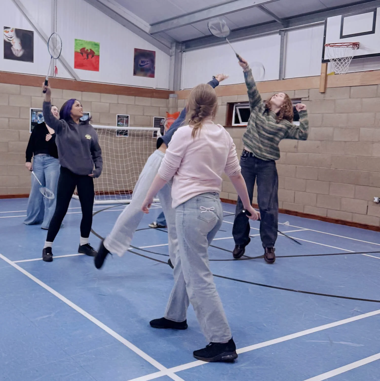 A group of young women playing badminton indoors on a blue court. Three women are actively reaching for the shuttlecock, while others watch. The setting appears to be a gymnasium with a basketball hoop and sports artwork on the wall.