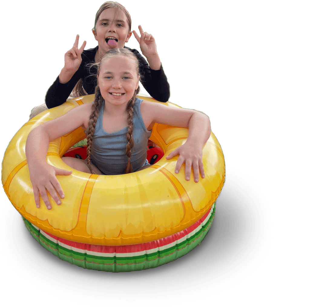 Two young girls sitting in a yellow and red inflatable swimming ring, smiling at the camera. The girl in front has long braided hair and is wearing a gray tank top. The girl behind has her tongue out and is making peace signs with both hands. The background is transparent.