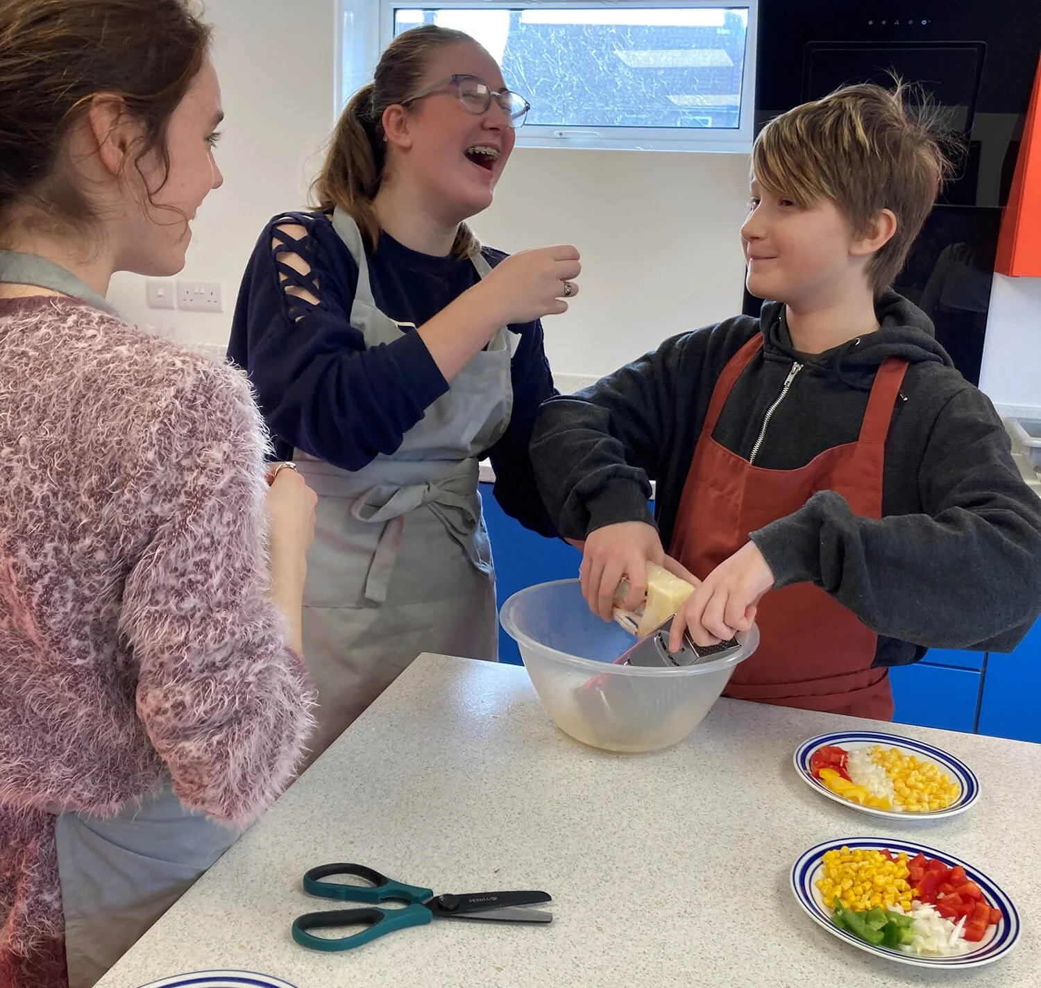 Three people in a kitchen preparing food, with plates of chopped vegetables and a bowl of salad ingredients on the counter, and a boy grating cheese into a plastic bowl.