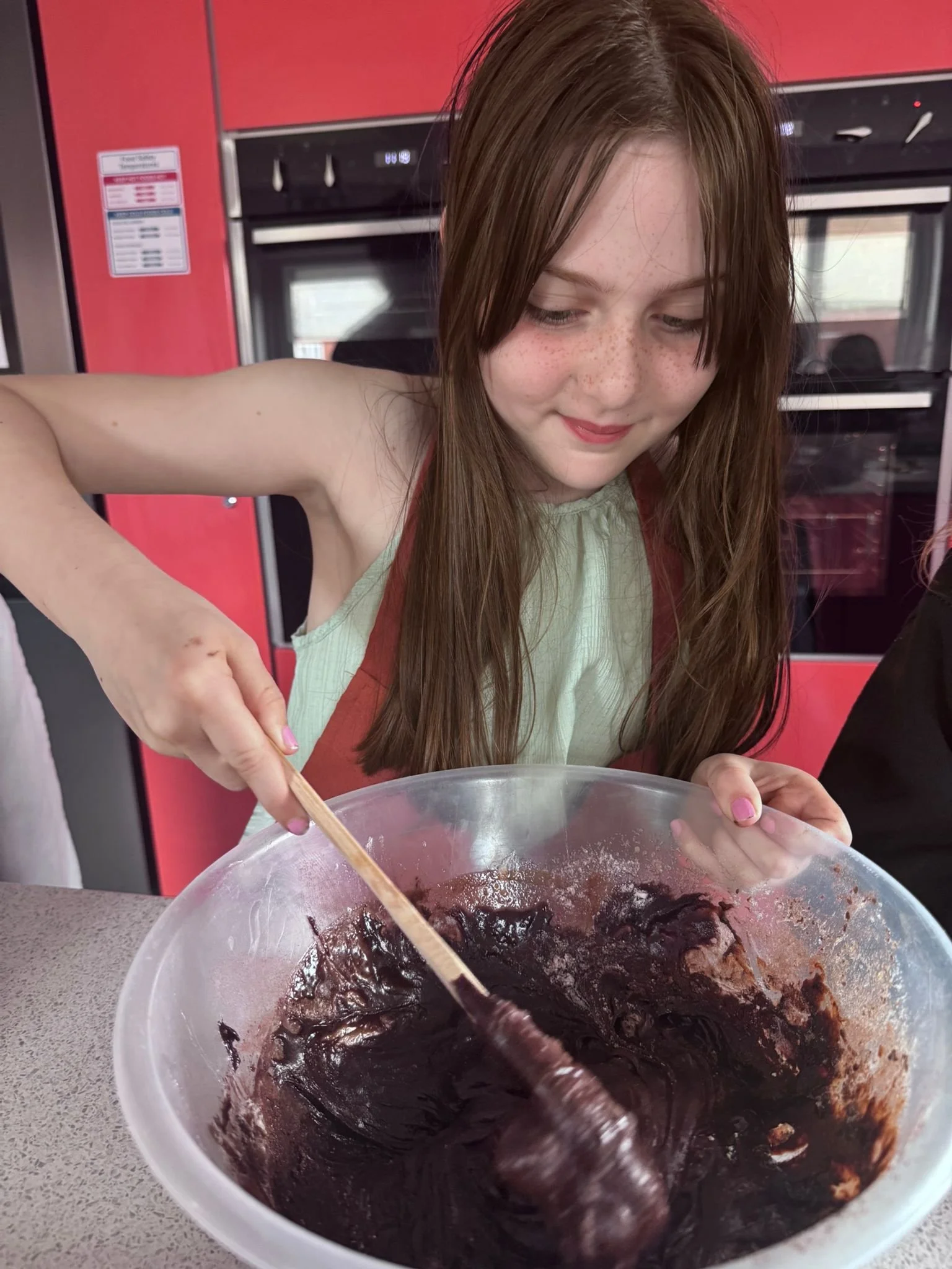 A young girl with long brown hair and freckles, stirring a large bowl of dark chocolate batter in a kitchen with red appliances.