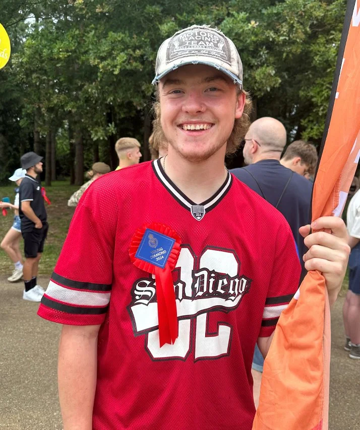 A young man smiling at a sports event, wearing a red jersey with the number 12 and a blue award ribbon, holding an orange flag, with a crowd and trees in the background.